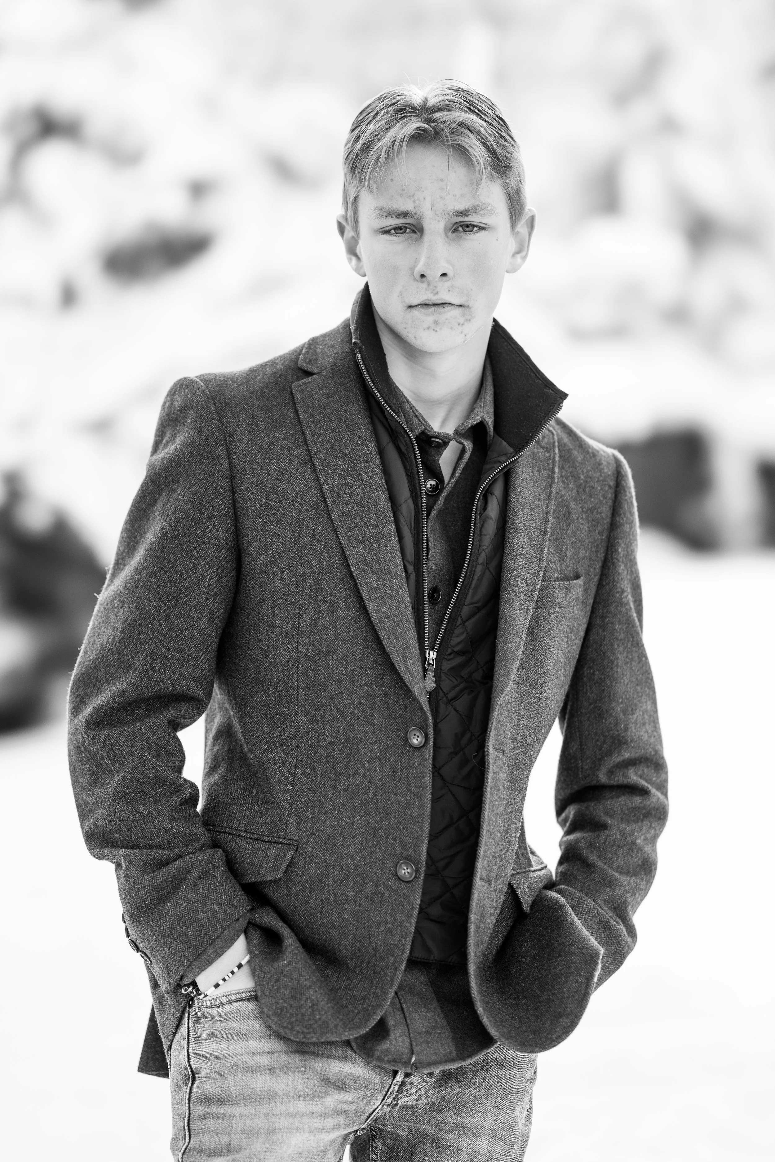A young man with light hair and freckles stands outdoors in winter, wearing a dark blazer over a quilted vest and a shirt. His hands are in his pockets, and he looks into the camera with a serious expression.