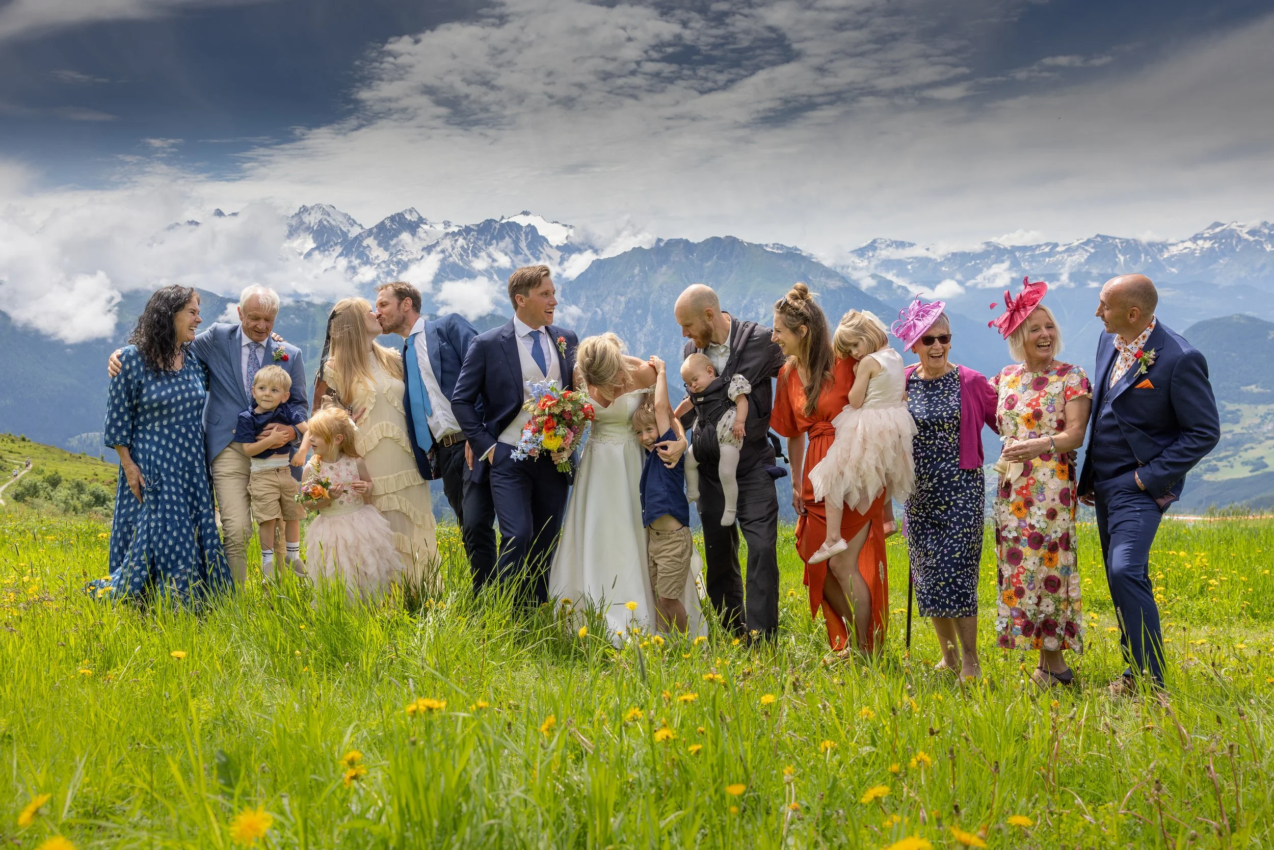 Group of people, including children, gathered outdoors in a grassy field with mountains and clouds in the background, celebrating a wedding at a wedding in Verbier Switzerland.