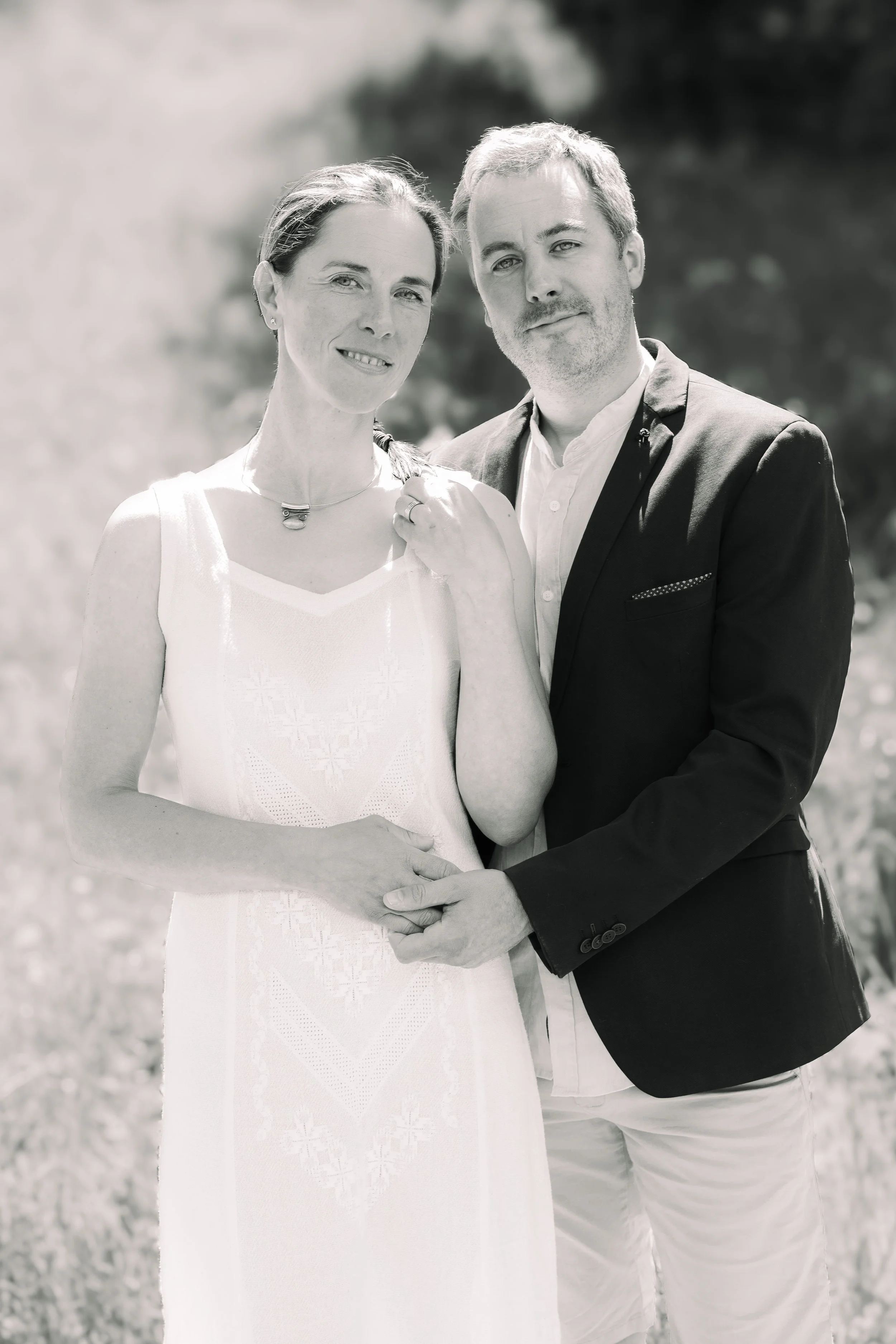 A black-and-white photo of a couple standing outdoors, holding hands and smiling at the camera. The woman is wearing a sleeveless white dress and a necklace, and the man is dressed in a dark blazer over a white shirt.