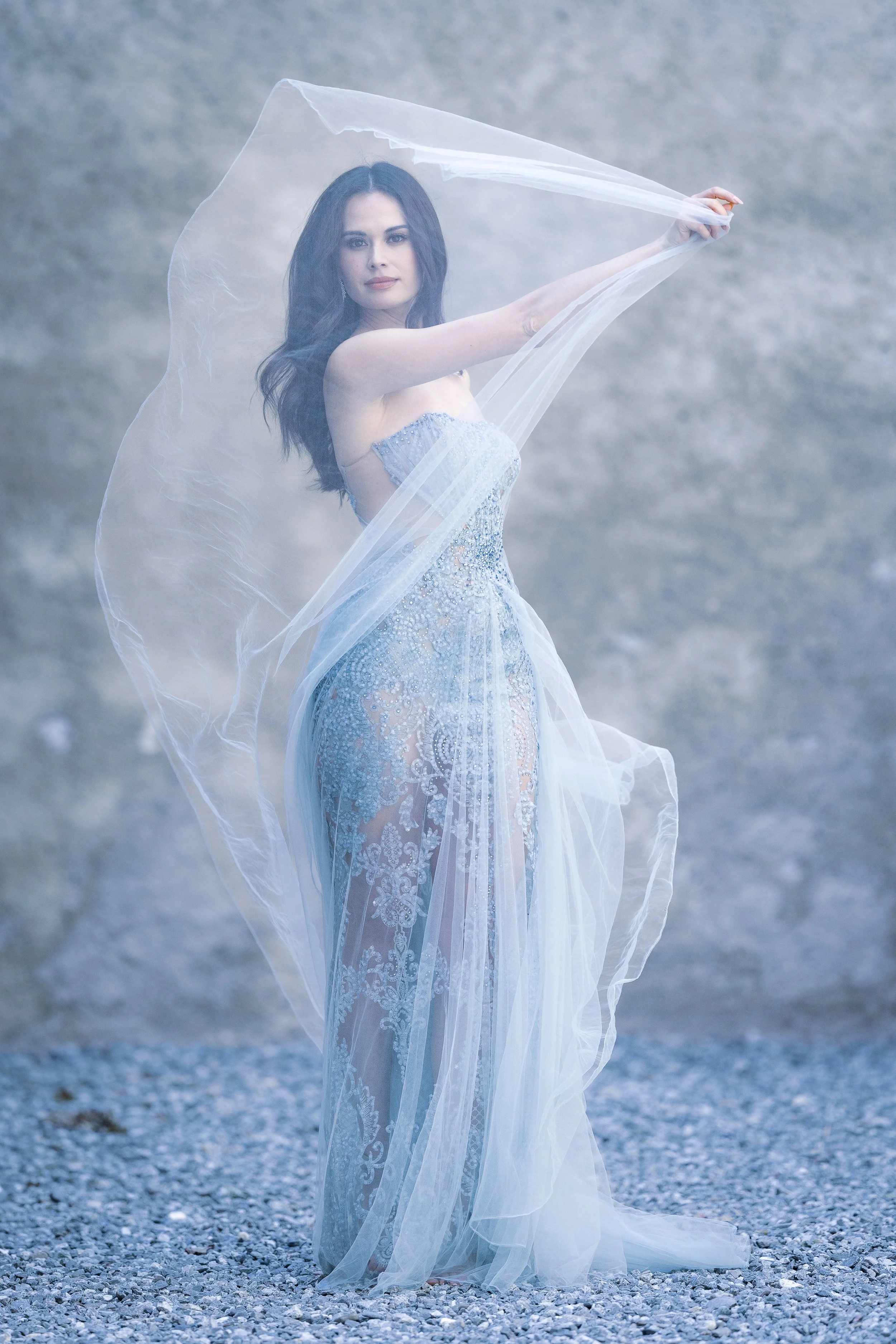 A woman in a sheer, light blue, lace gown standing outdoors on a gravel surface, holding a transparent fabric above her head with both hands, against a gray, cloudy background.