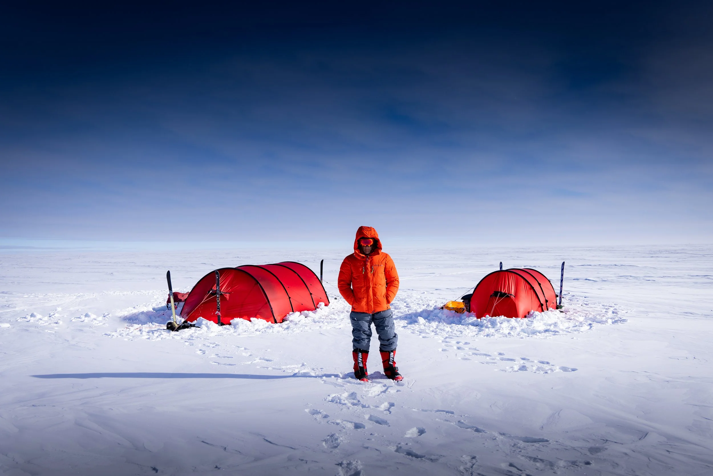 A person in Arctic gear standing on snow with two red tents and snow shovels nearby under a clear blue sky.
