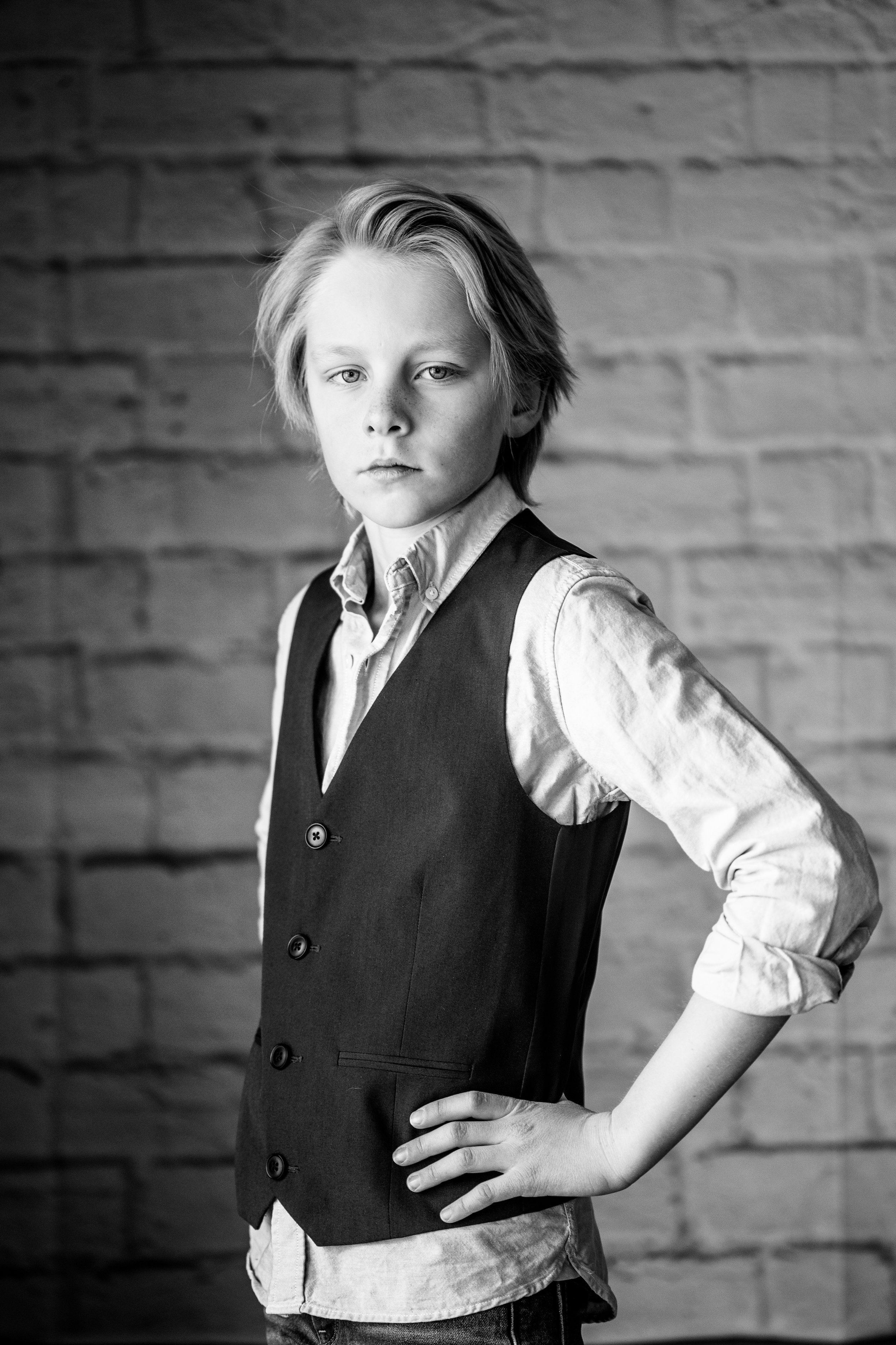 A young boy with shoulder-length hair standing with one hand on his hip, wearing a light-colored shirt and a dark vest, against a brick wall background.