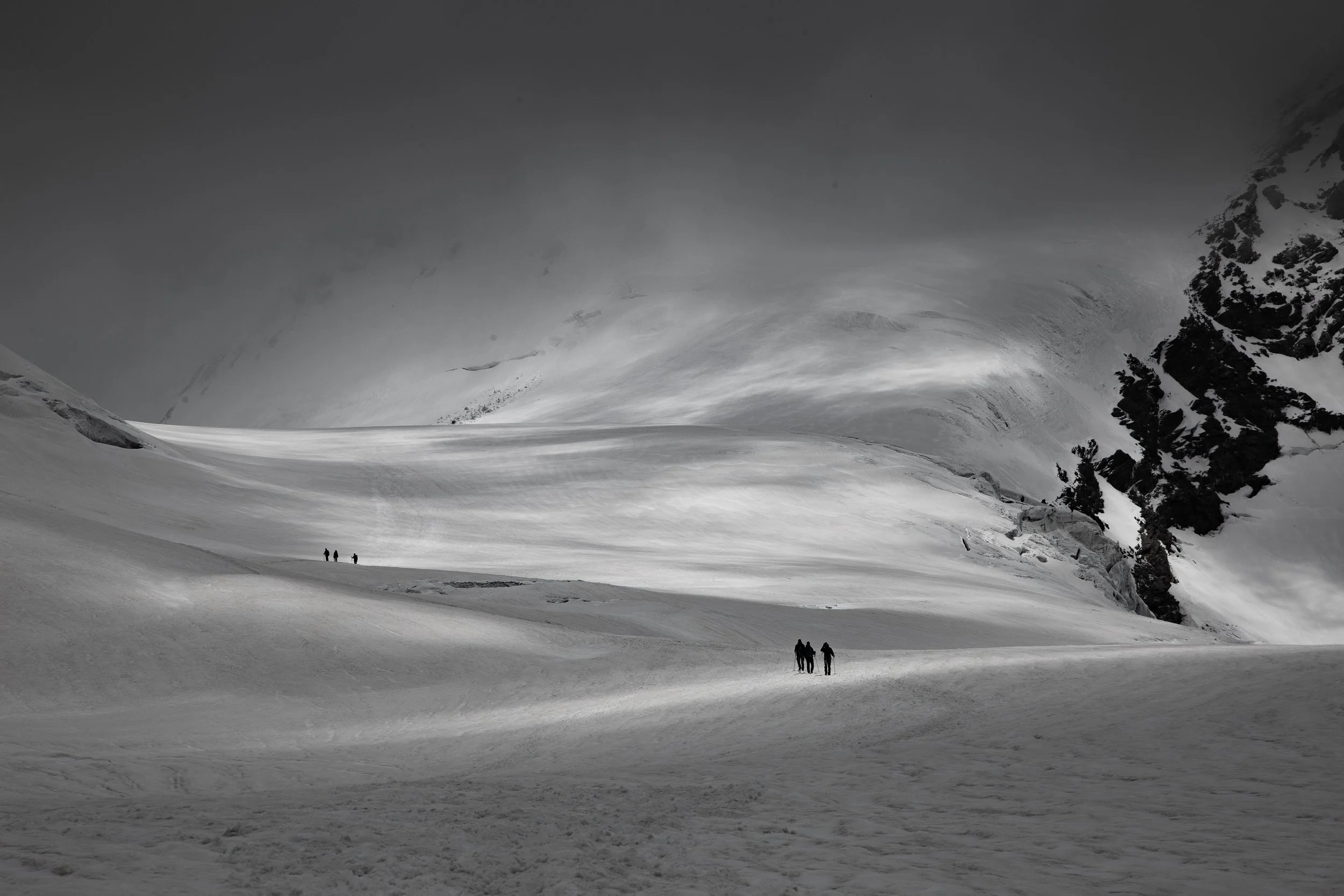 Four people walking across a snow-covered mountain landscape, with steep slopes and snow-covered rocks, under a cloudy sky.