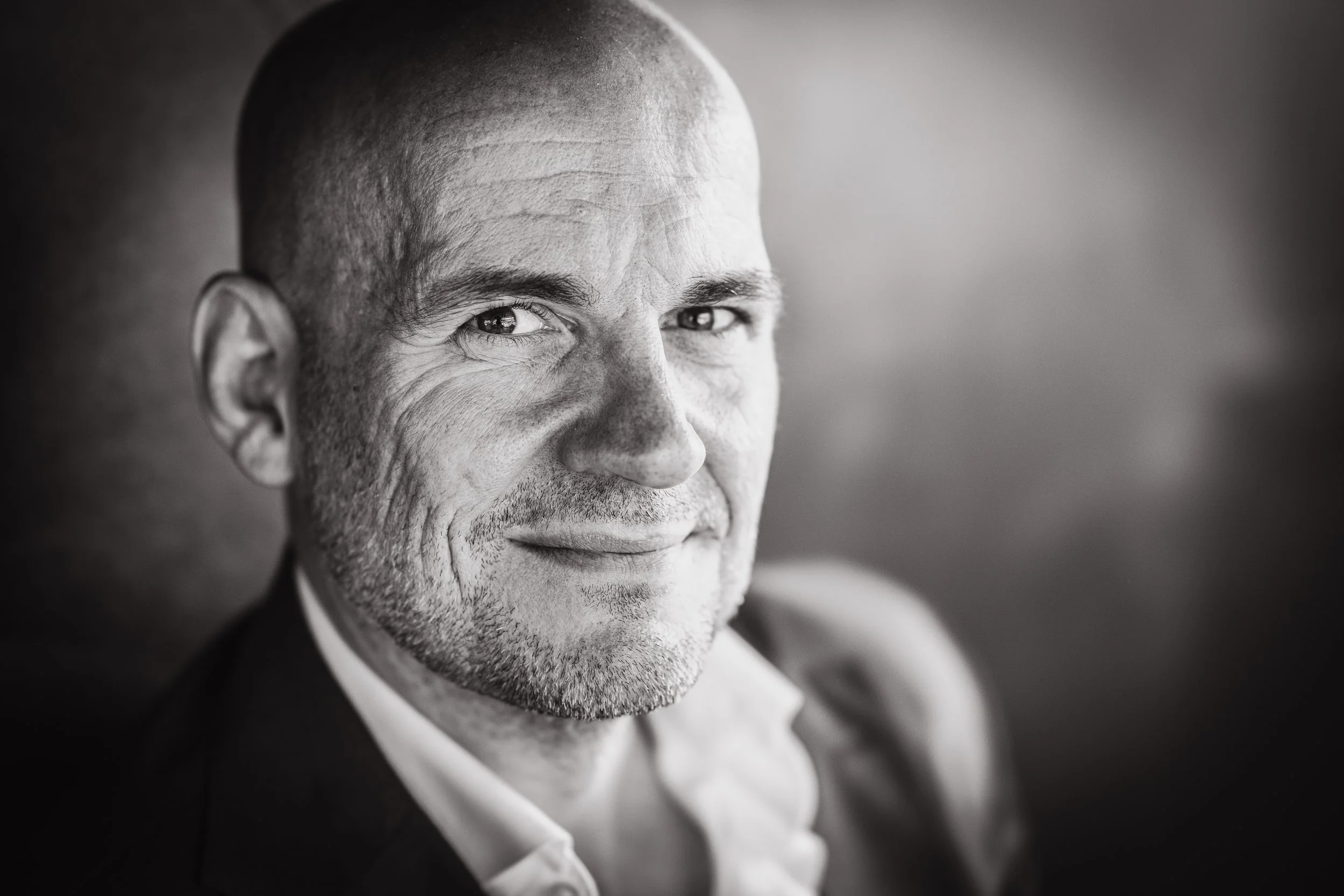 Black and white close-up portrait of a smiling middle-aged man with a shaved head, wearing a collared shirt and blazer, looking slightly to the left.