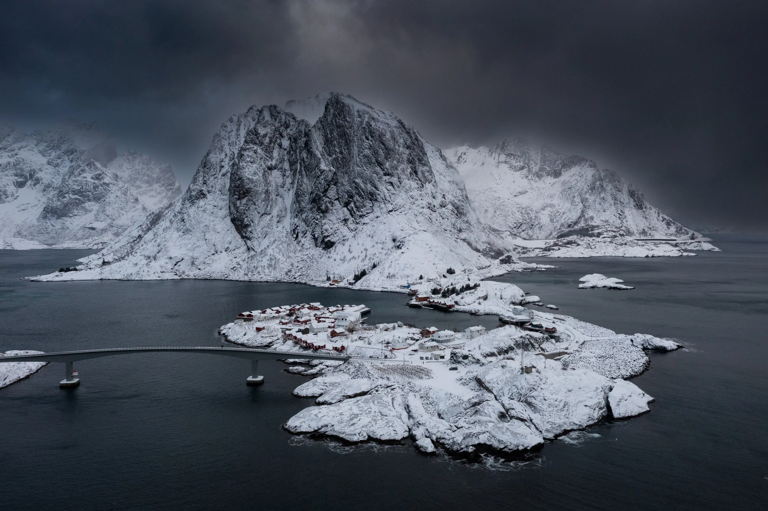 A snowy island with a small village and a bridge, surrounded by water and tall snow-covered mountains under a dark, cloudy sky.