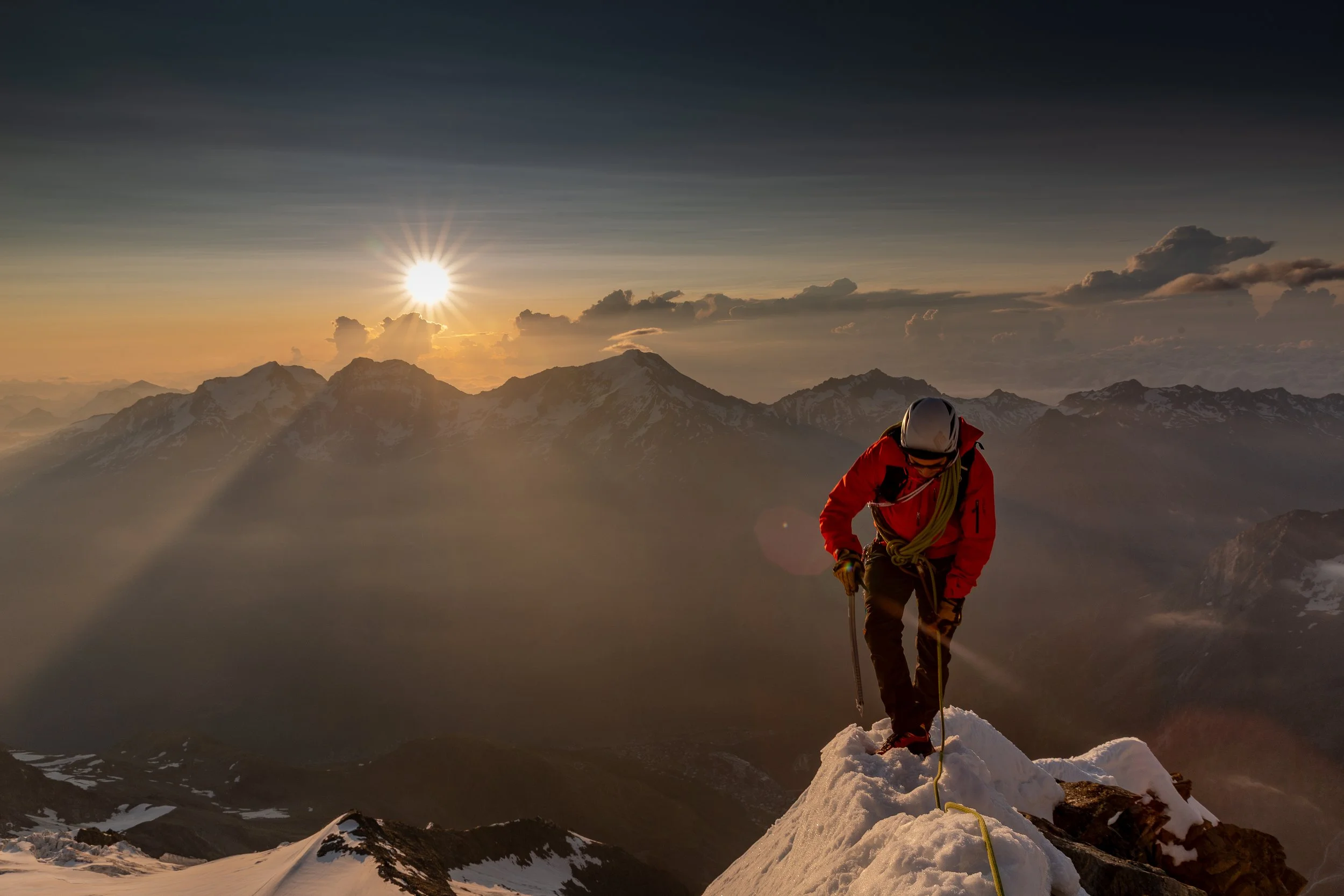A mountaineer in a red jacket and helmet climbing a snow-covered peak during sunset in the mountains.