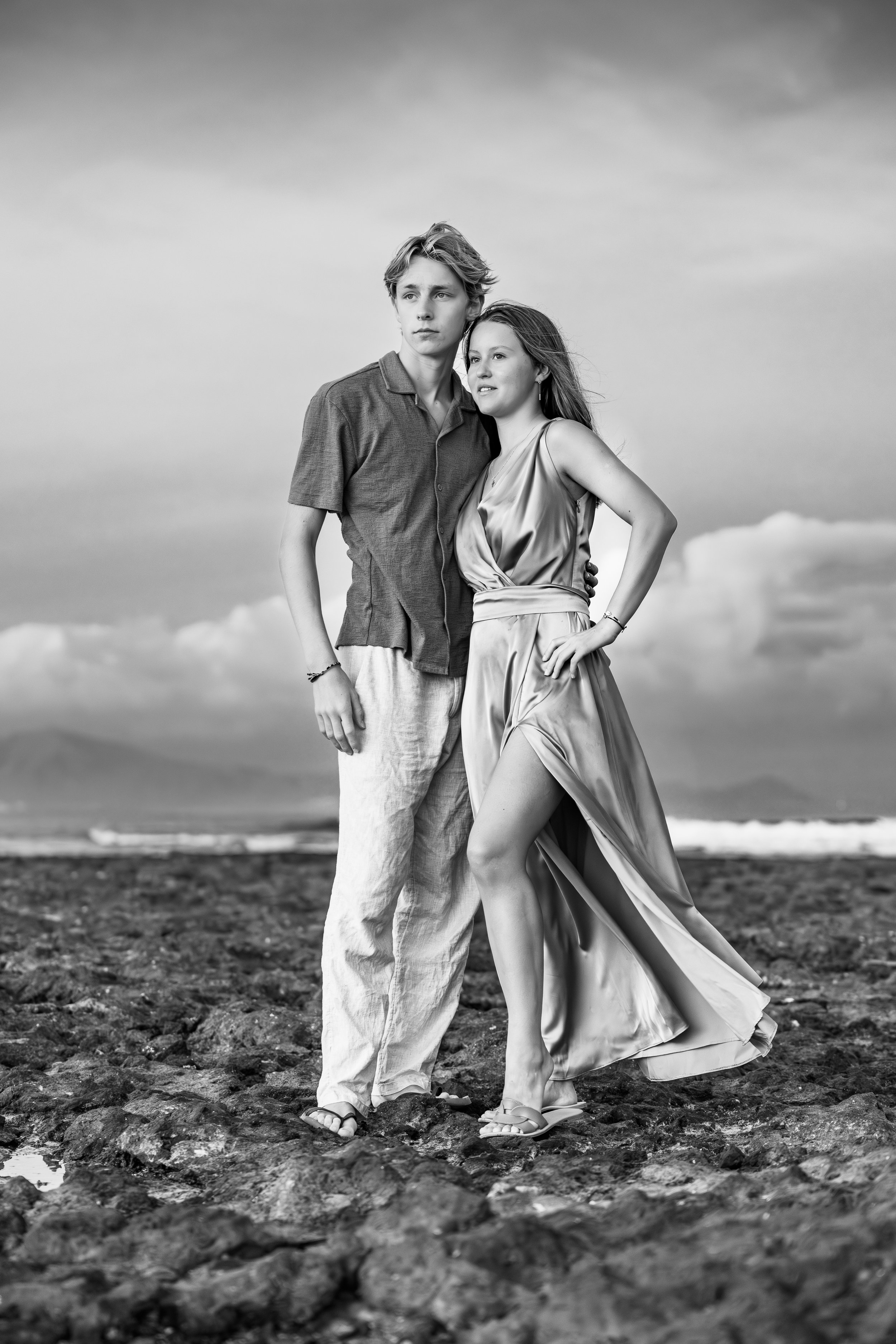 Young man and woman standing on a rocky beach, the woman is wearing a flowing dress, both looking into the distance, with the ocean and cloudy sky in the background.