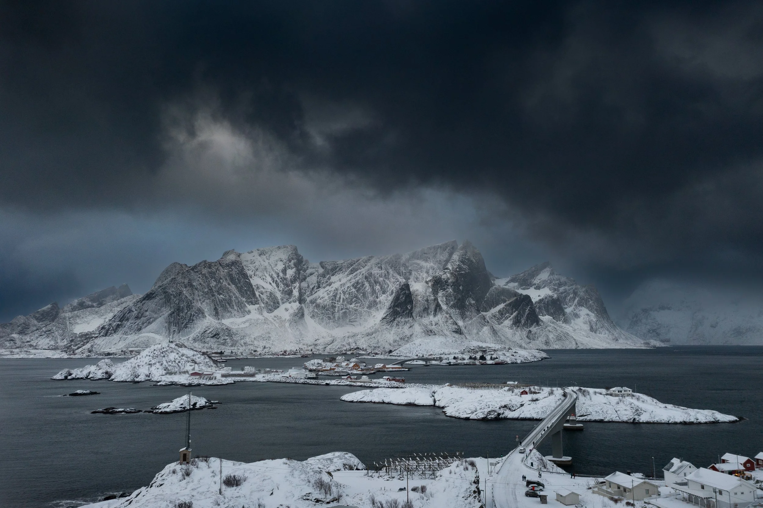Snow-covered island with a bridge, buildings, and mountains under dark, cloudy skies in a winter landscape.