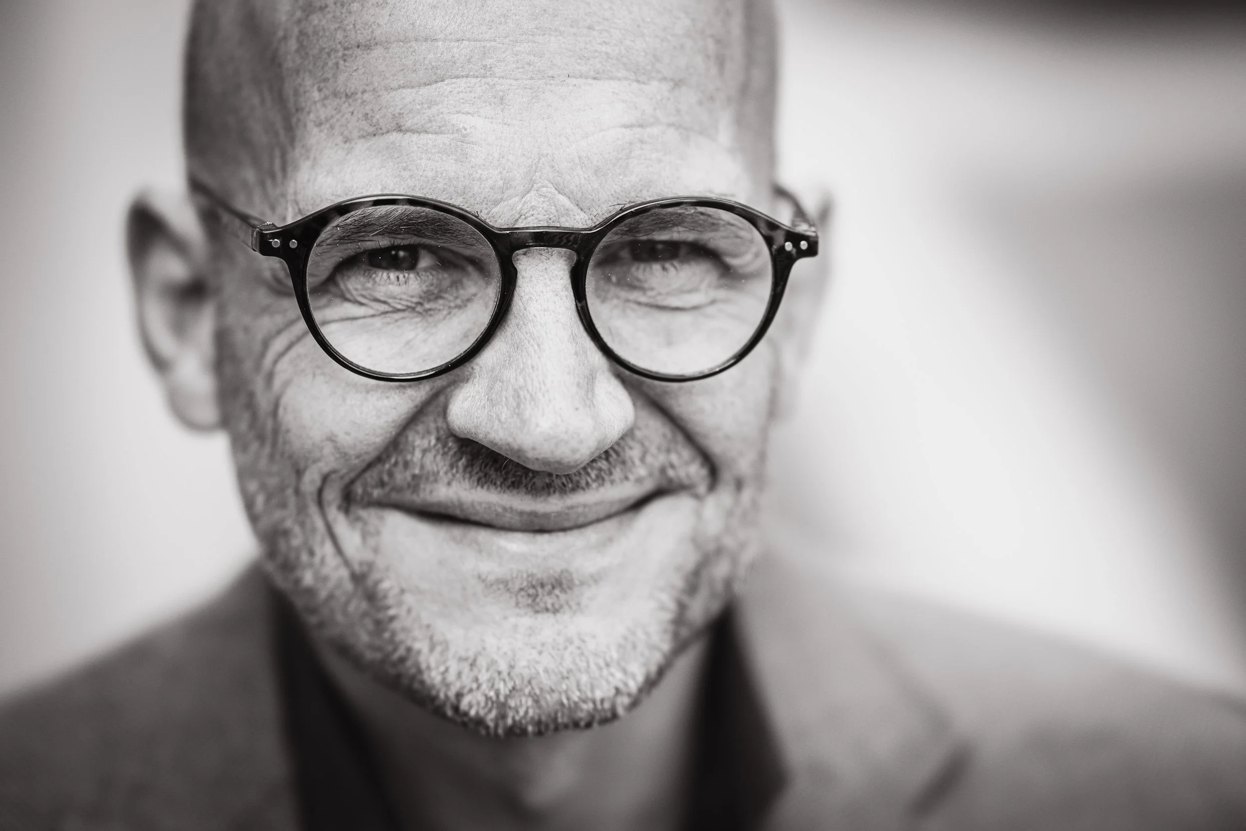 Close-up black and white photo of a smiling middle-aged man with glasses, showing wrinkles and stubble, wearing a suit.