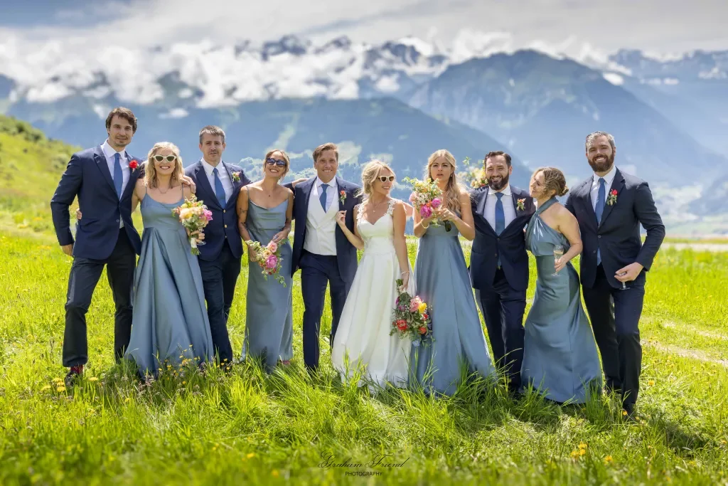 A wedding party of ten people, including the bride in a white dress and nine bridesmaids and groomsmen in blue dresses and suits, standing together on a grassy field with mountains in the background at a wedding in Verbier Switzerland.