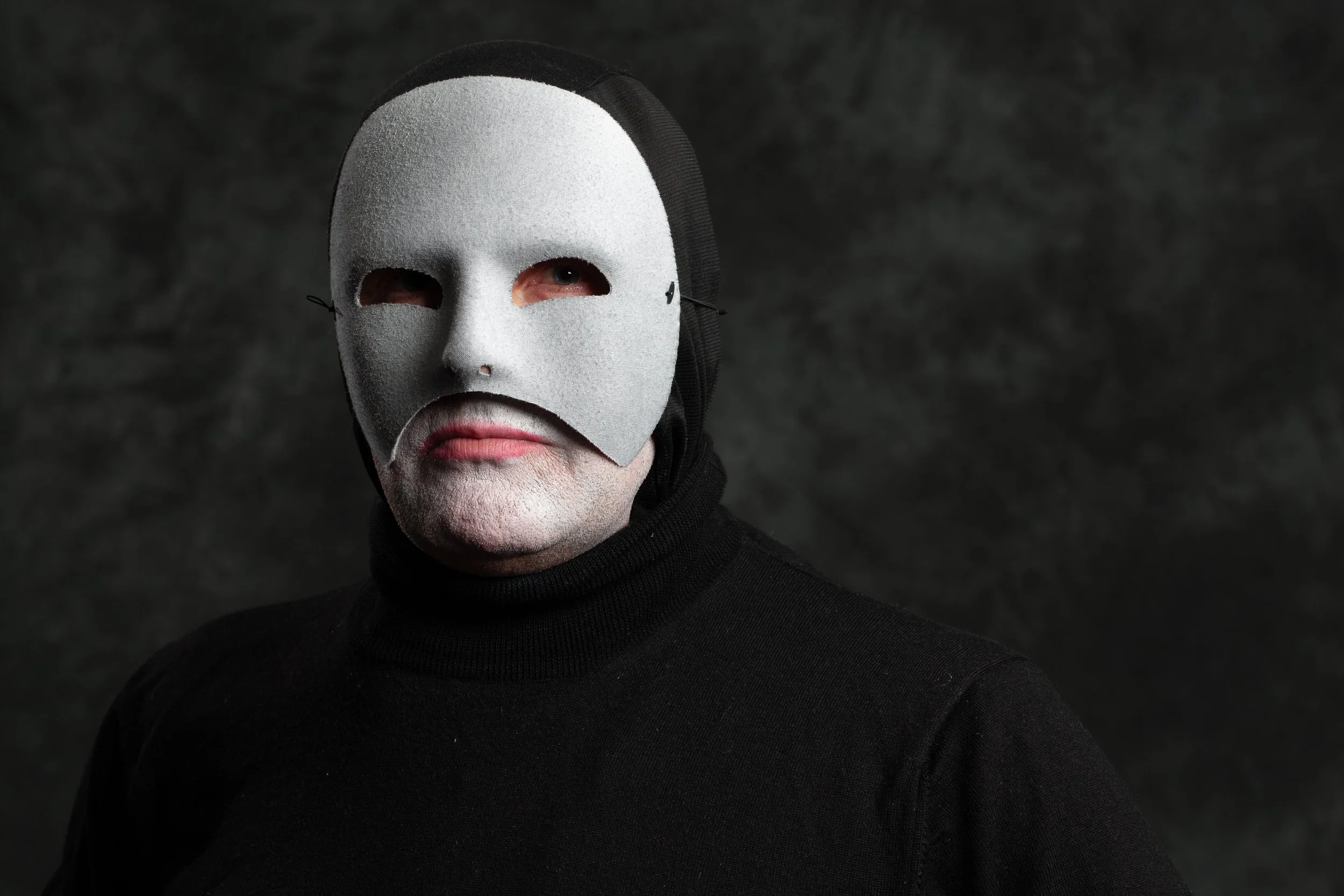 A person wearing a black turtleneck and a white mask with eye and mouth holes, with a dark textured background.