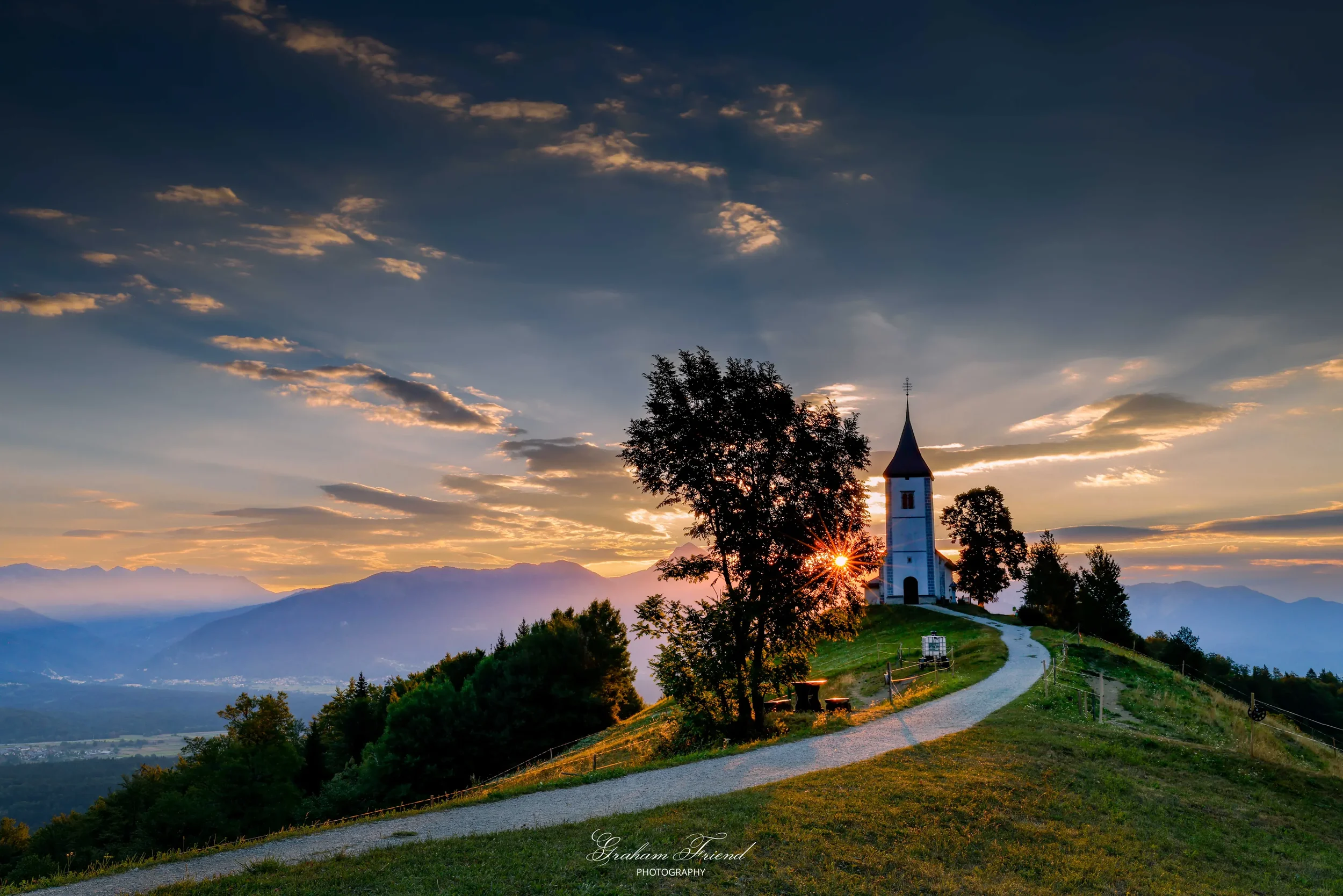 A scenic view of a small white church on a hill at sunset, with a dirt path leading up to it, surrounded by trees and mountains in the distance.
