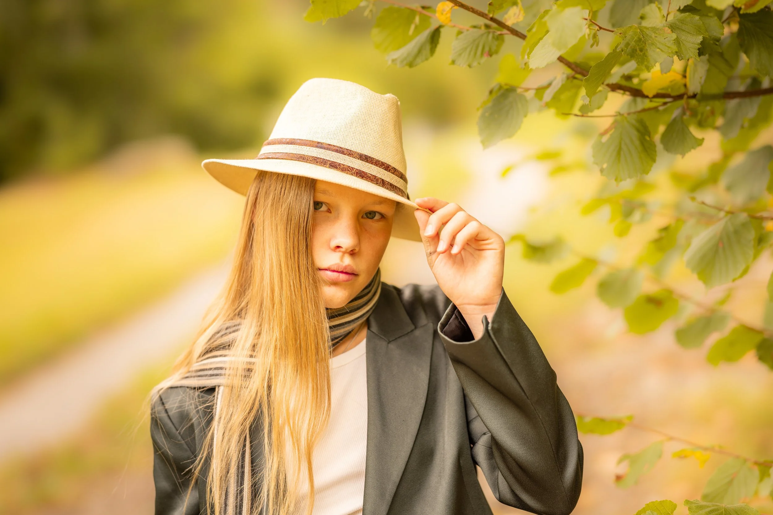 A young woman with long red hair wearing a beige hat, black blazer, and striped scarf, standing outdoors among green leaves.