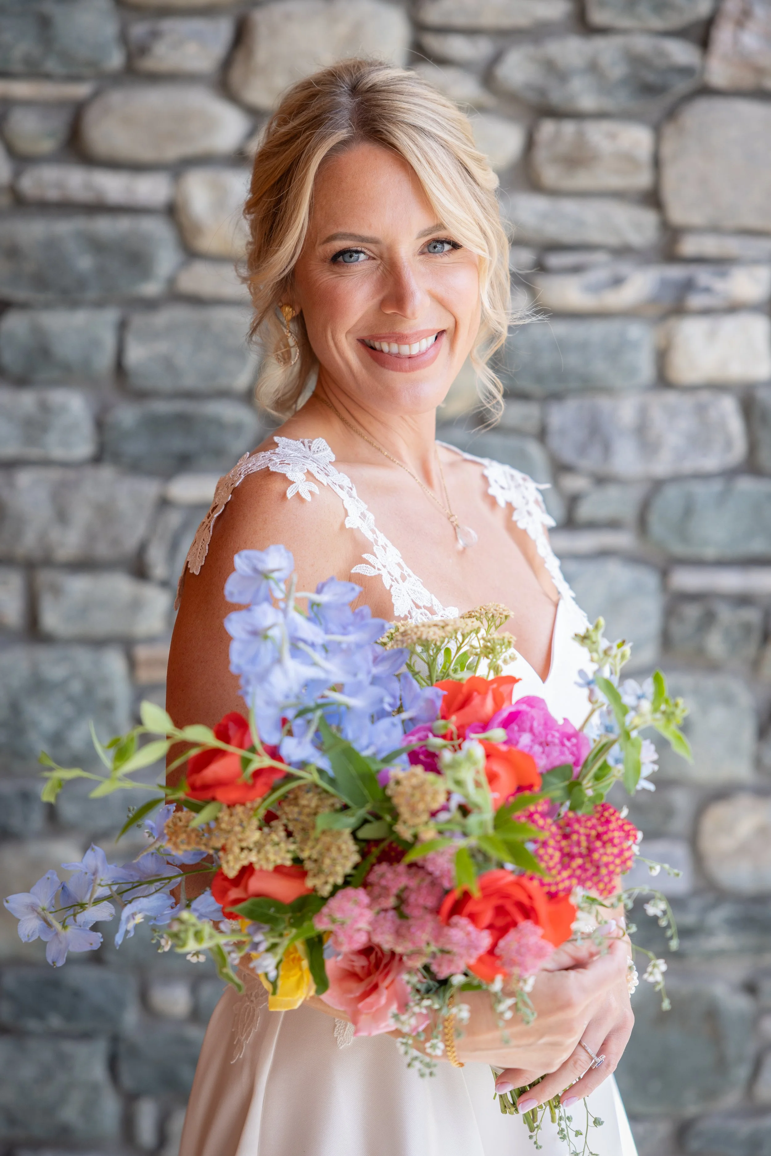 A smiling bride from a wedding in  with blonde hair styled in loose waves, wearing a white wedding dress with lace shoulder details, holding a colorful bouquet of mixed flowers including pink, orange, purple, and green hues, in front of a stone wall.