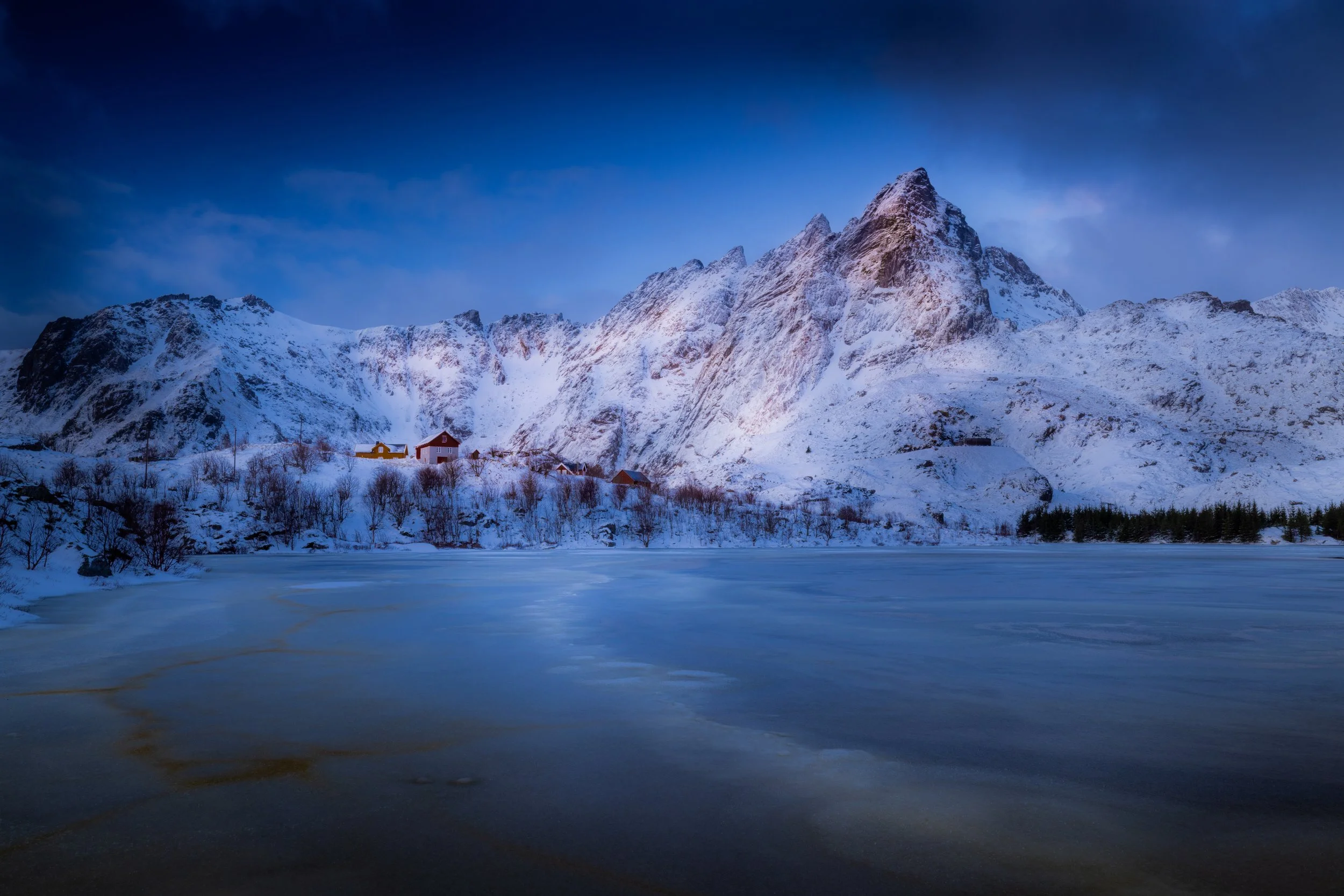 Snow-covered mountains beside a frozen lake with small houses on the shore under a cloudy sky.
