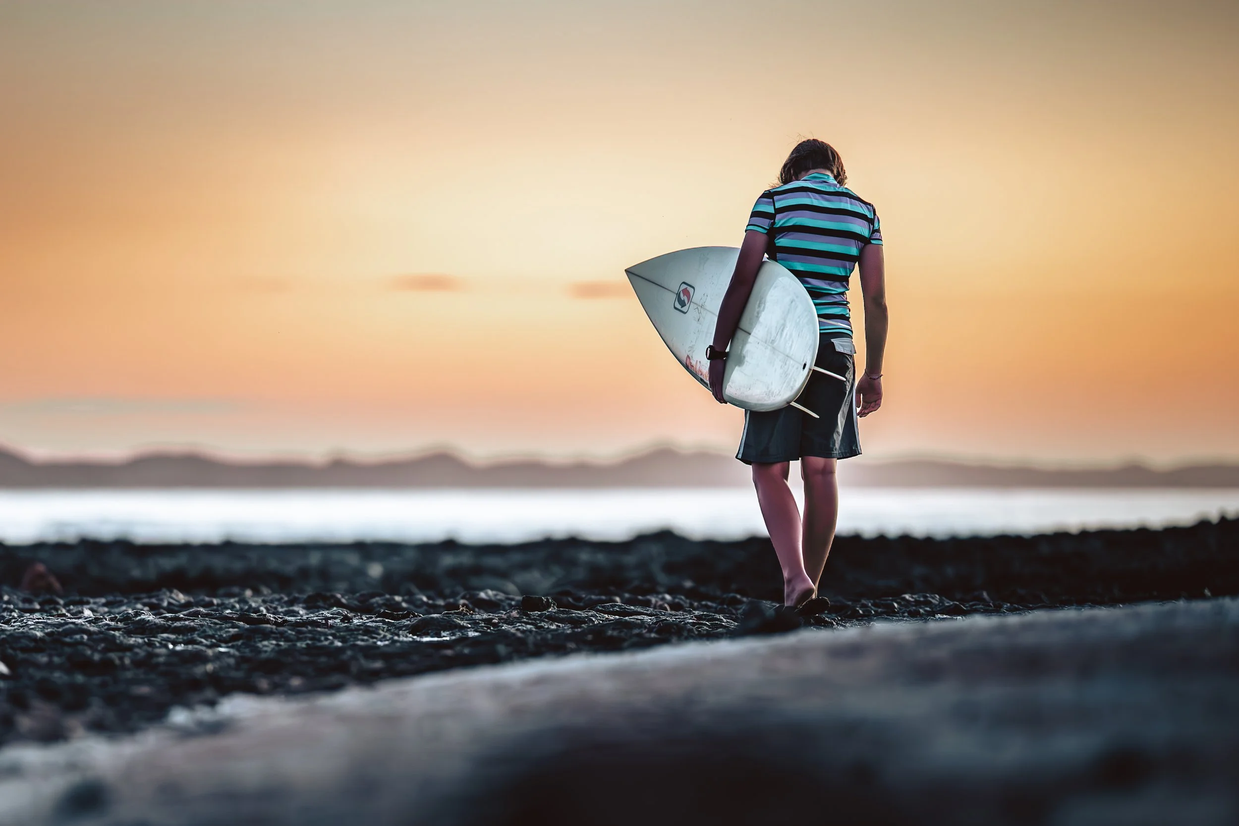 A person walking on a rocky beach at sunset, holding a surfboard under their arm, wearing a striped shirt and shorts.