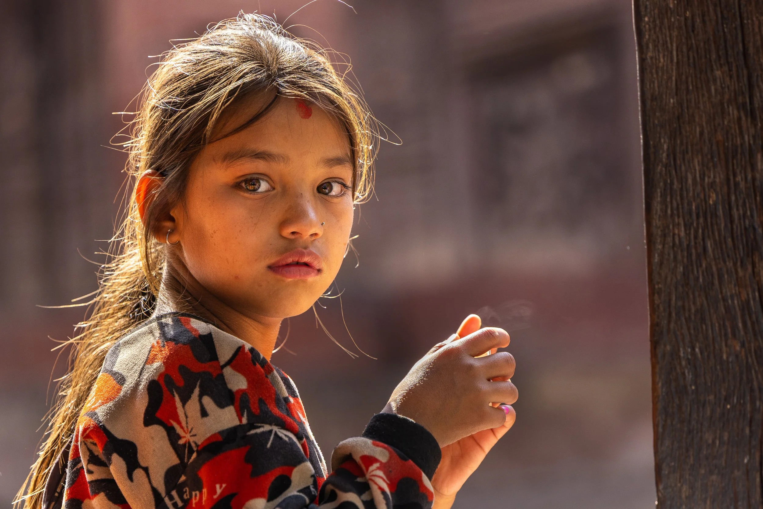 A young girl with brown hair and brown eyes looking towards the camera, holding a wooden pole. She is wearing a colorful patterned shirt and has a small nose piercing.