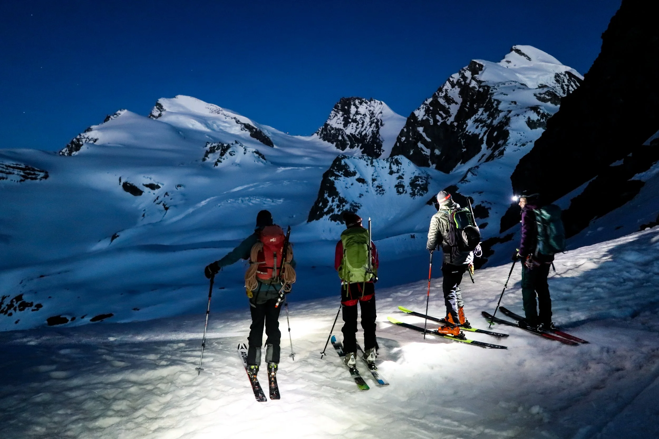 Four hikers with backpacks and snowshoes stand on snowy terrain at night, looking at snow-covered mountains under a dark sky.