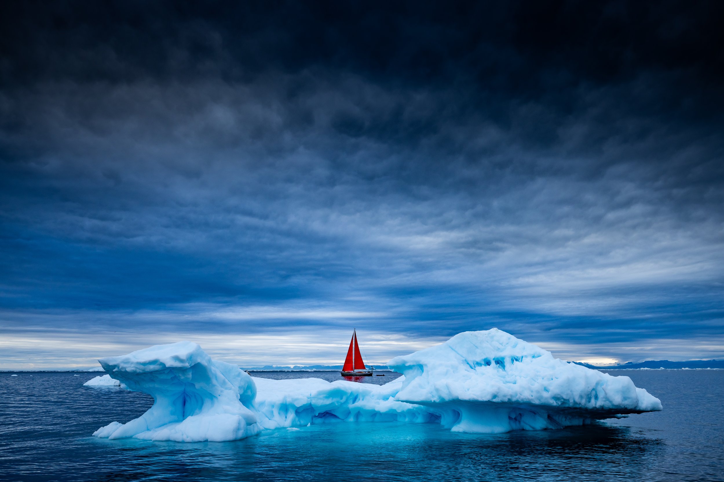 An iceberg floating in Arctic waters with a red-sailed boat in the background under a cloudy sky.