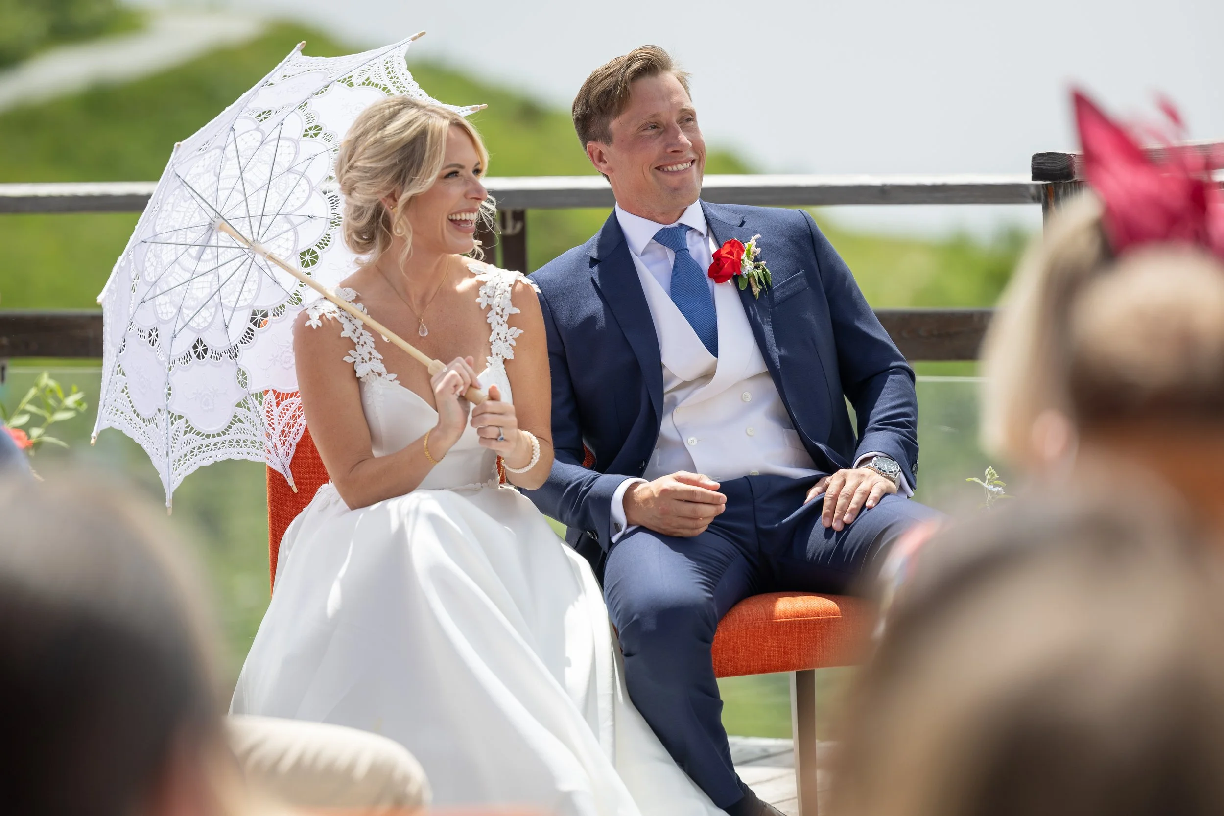 A bride and groom sitting outdoors at their wedding ceremony, smiling and enjoying the moment. The bride holds a lace parasol and wears a white wedding dress with lace details, while the groom is dressed in a navy blue suit with a red boutonniere at 