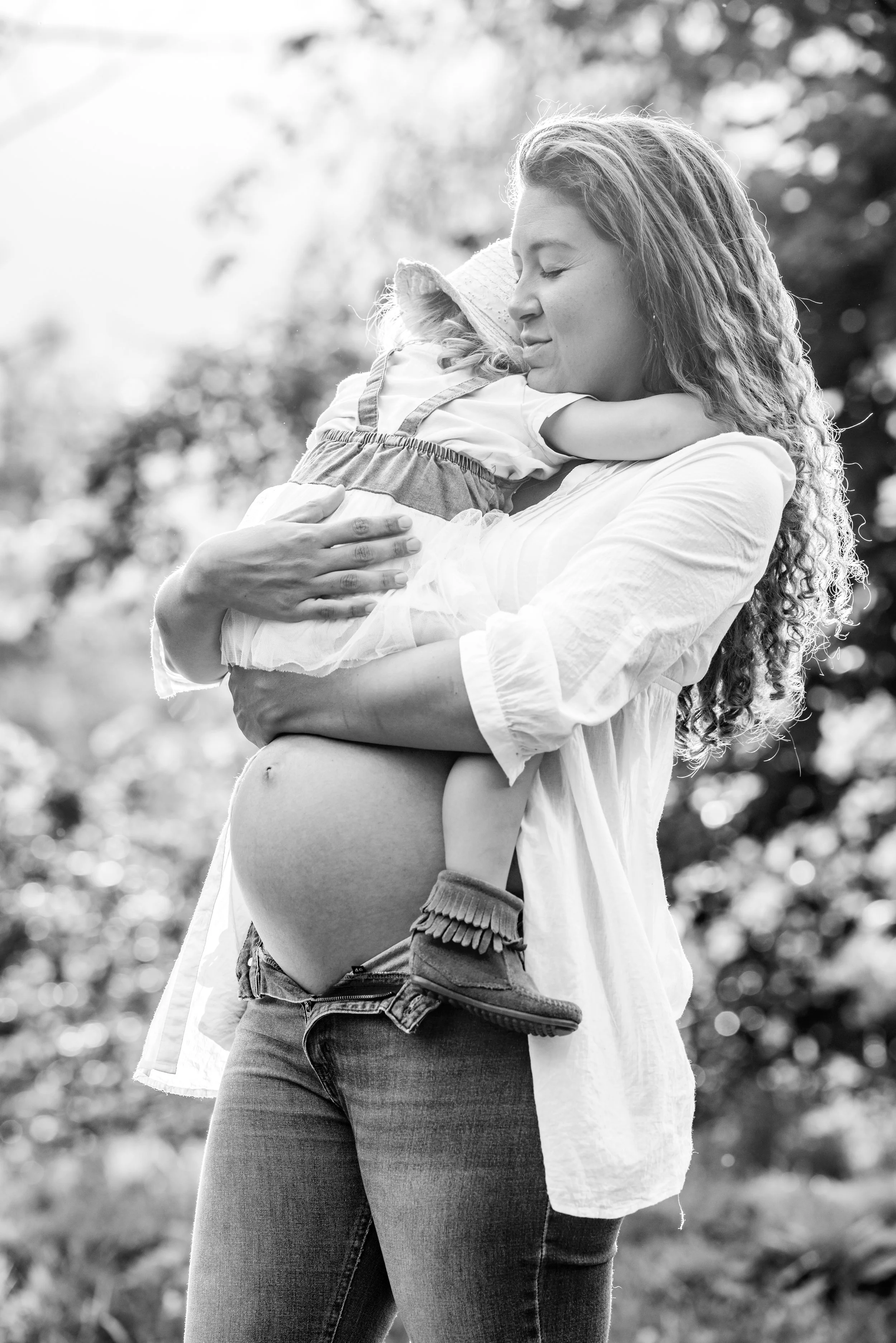 A woman with curly hair holding a young girl in her arms, both with closed eyes, outdoors in natural light.