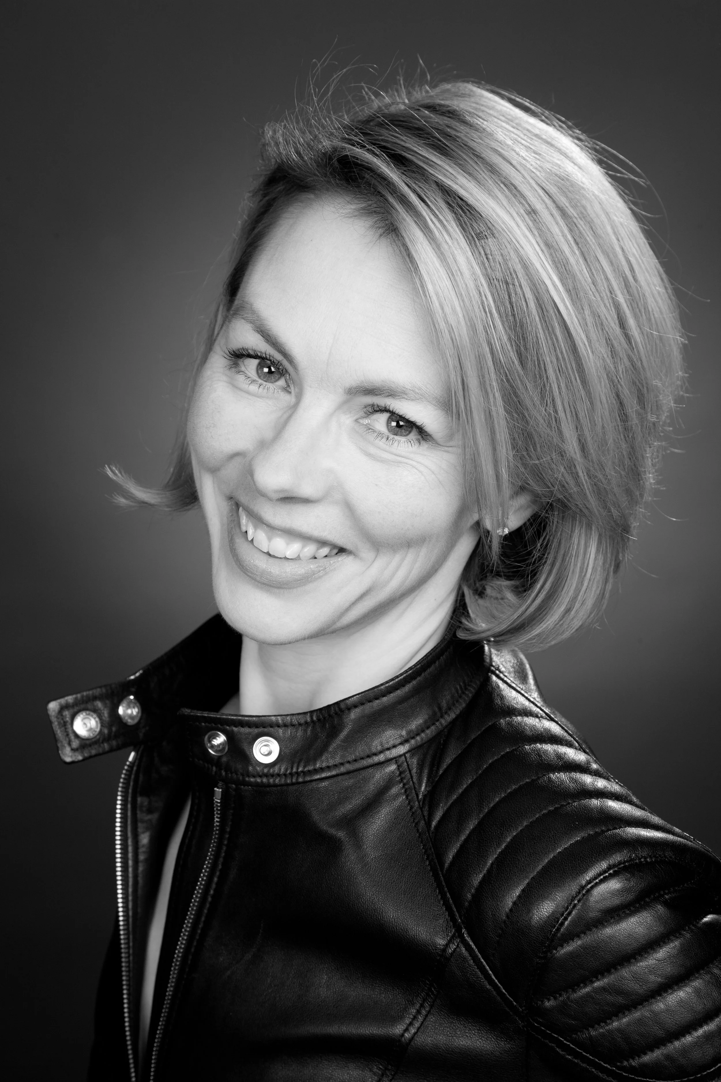Black and white portrait of a smiling woman with short hair wearing a leather jacket.