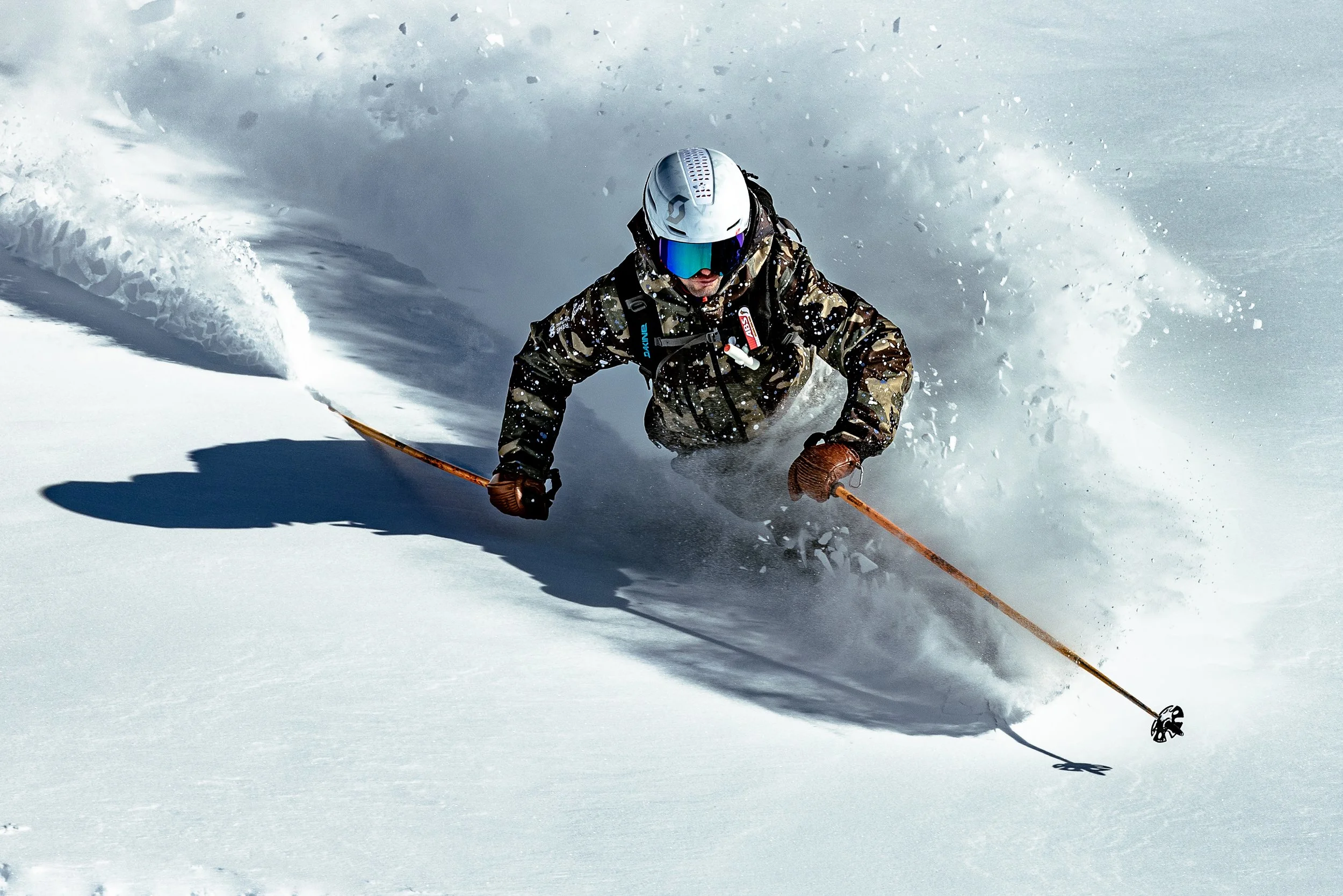 A skier in camouflage jacket and brown gloves skiing downhill through snow, wearing a white helmet and reflective goggles.