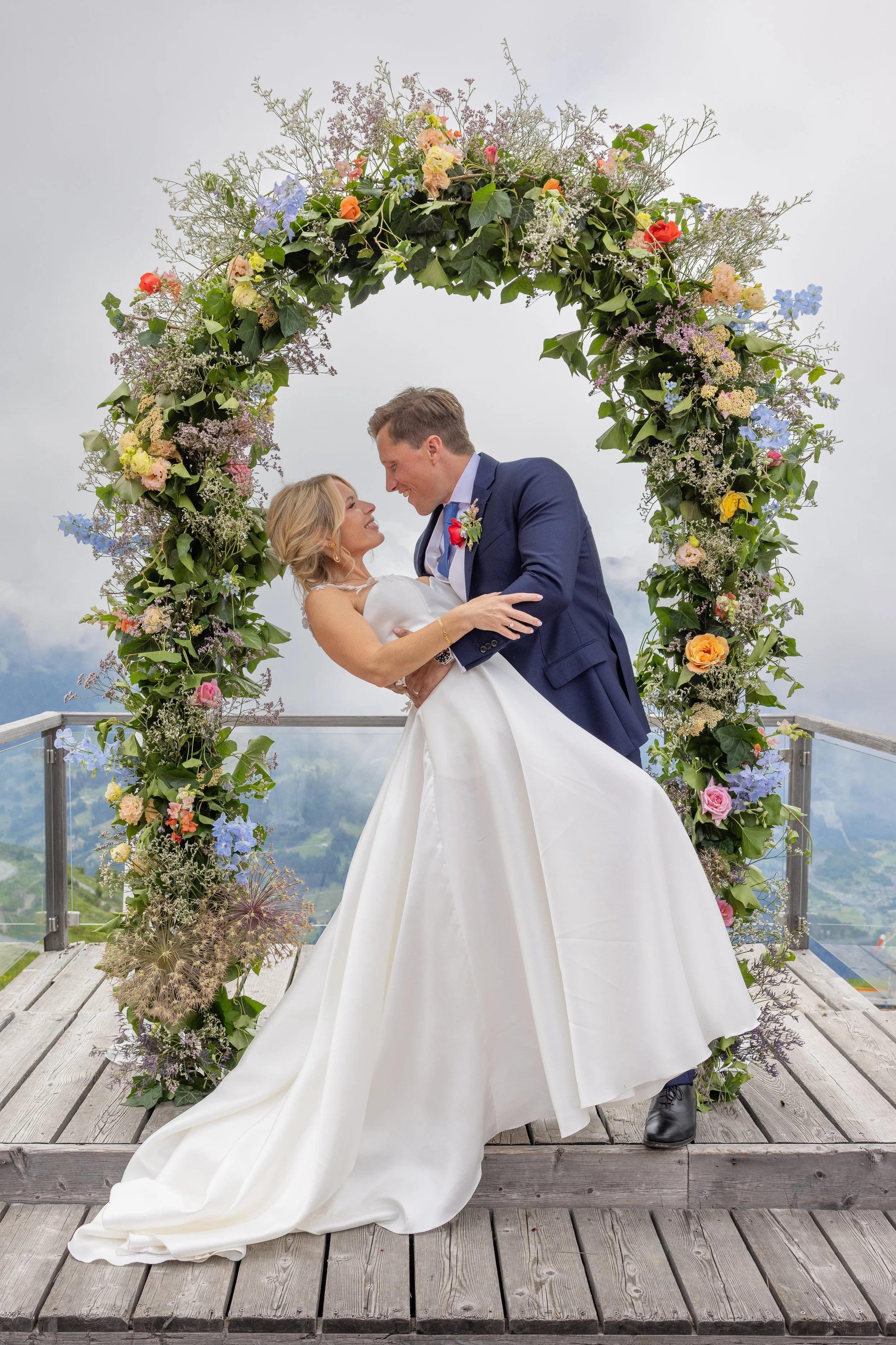 A bride and groom dance beneath a colorful floral arch on a wooden deck with a mountain view in the background at a wedding in Verbier Switzerland.