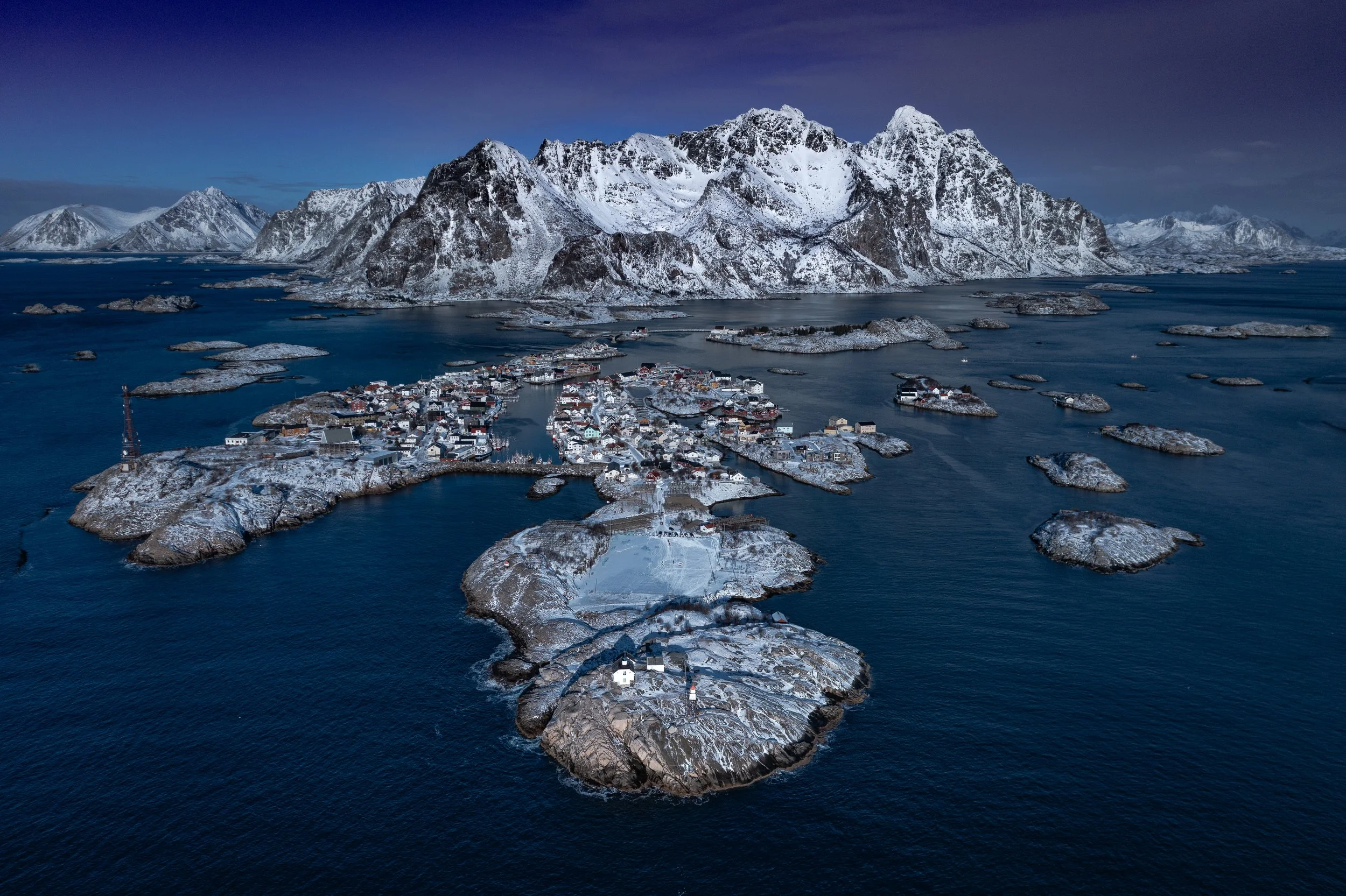 A snowy coastal island village with surrounding water and mountainous landscape in the background.