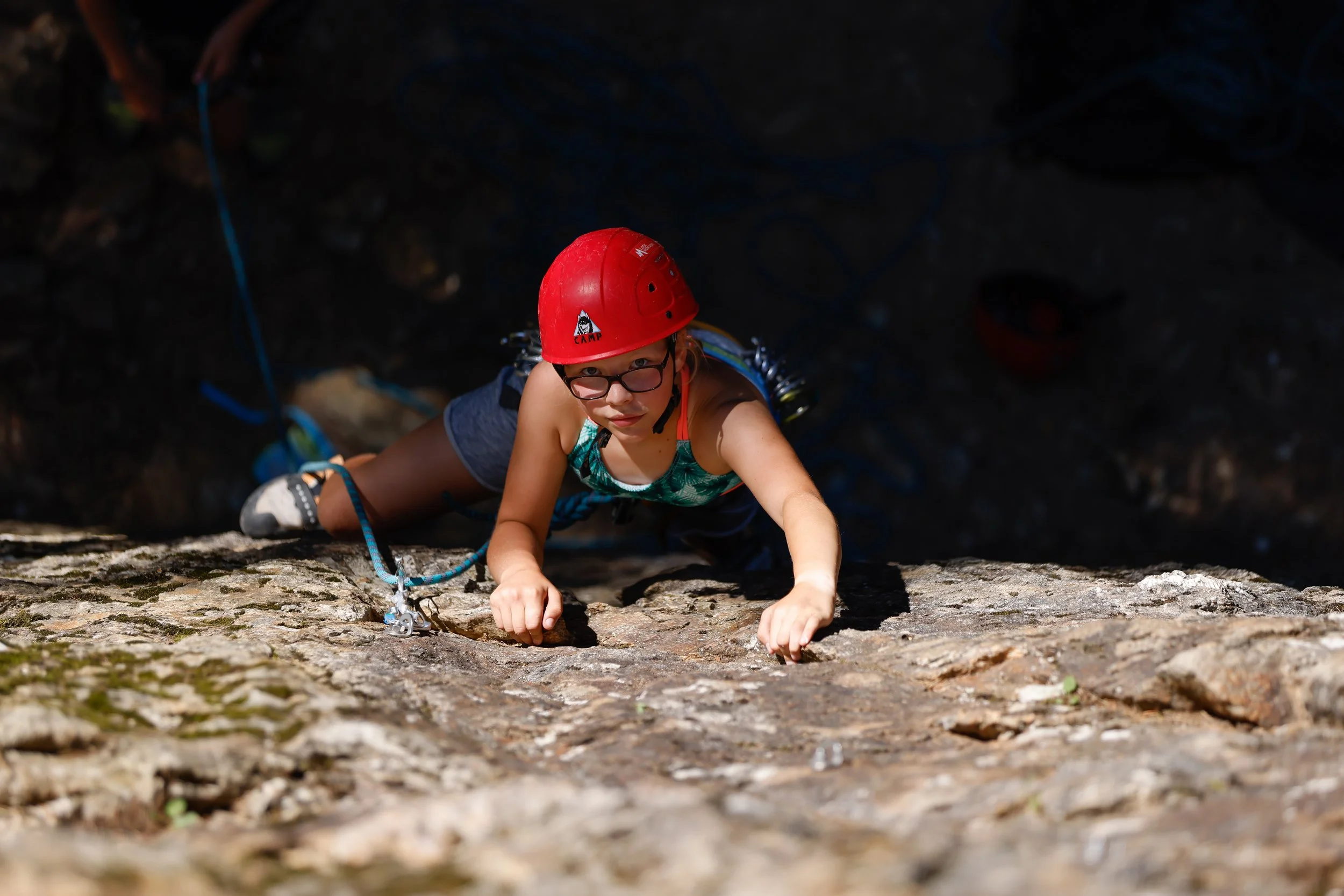 Young girl rock climbing outdoors, wearing a red helmet and glasses, gripping a rocky surface.
