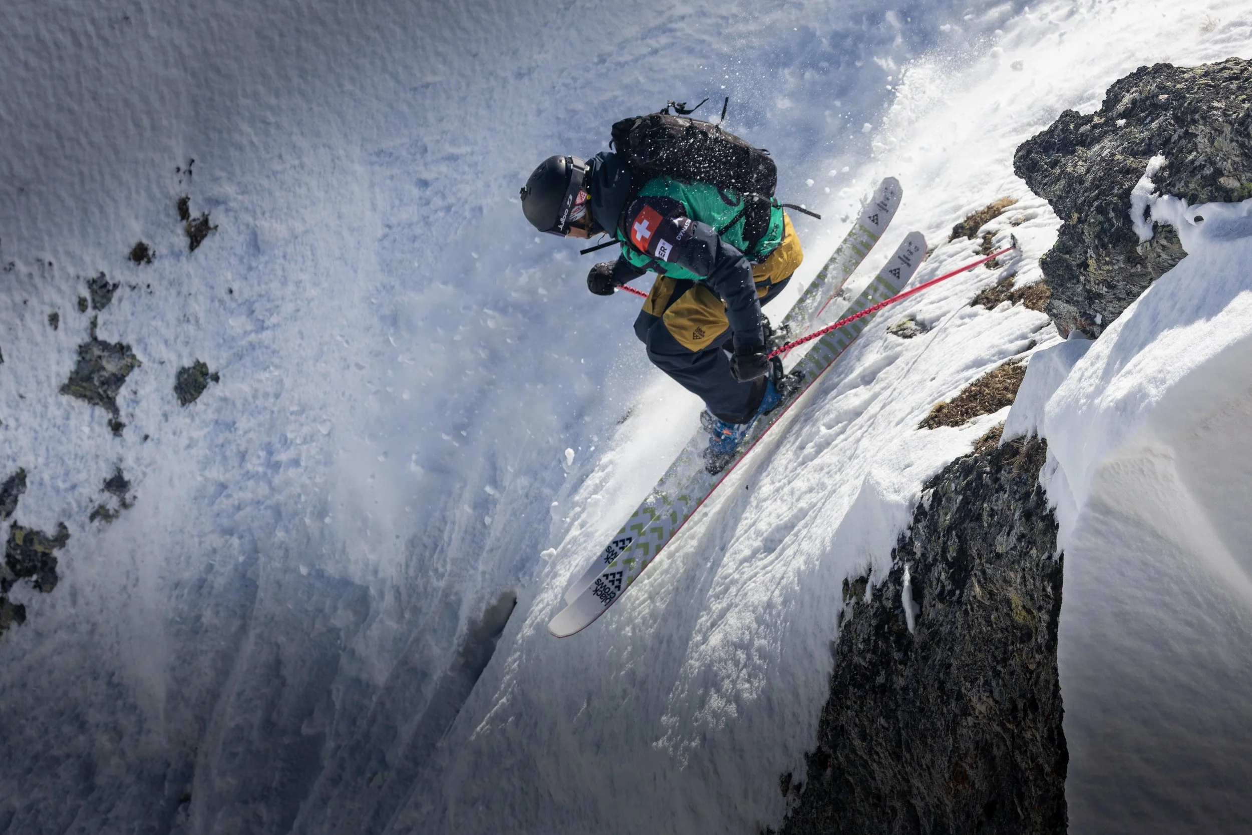 A skier in a green and yellow outfit with a Swiss flag patch on their sleeve descends a steep, snowy mountain slope with rocks, kicking up snow and ice.