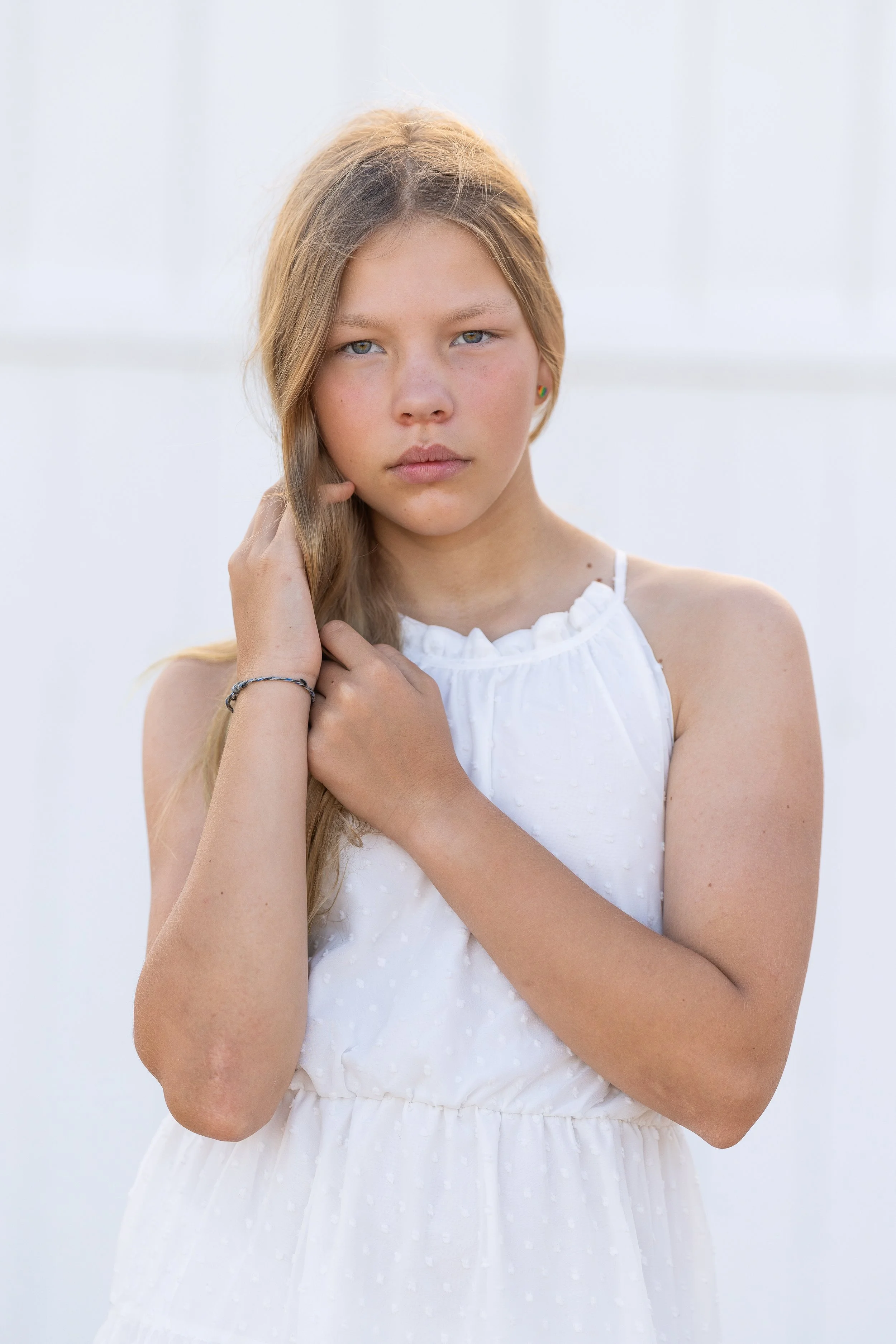 A young girl with long blonde hair and blue eyes wearing a white sleeveless dress, standing against a light background.