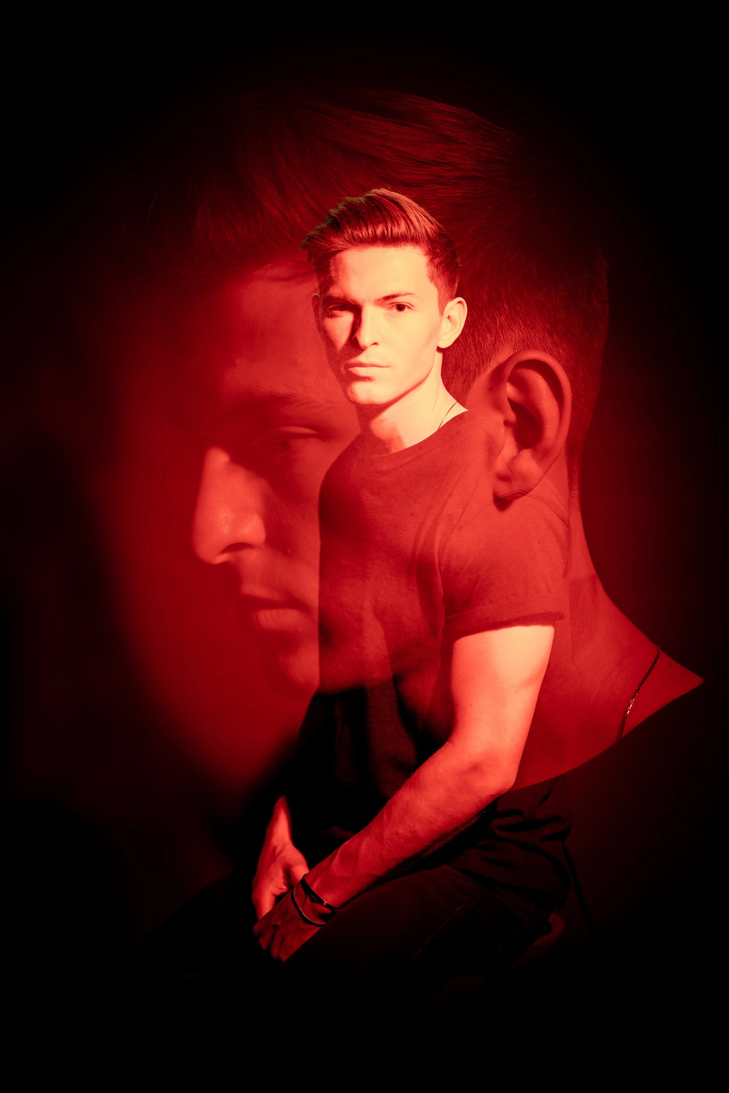 A digitally edited photo of a young man with blonde hair, wearing a black t-shirt, with a double exposure effect showing his face and upper body in red, against a black background.