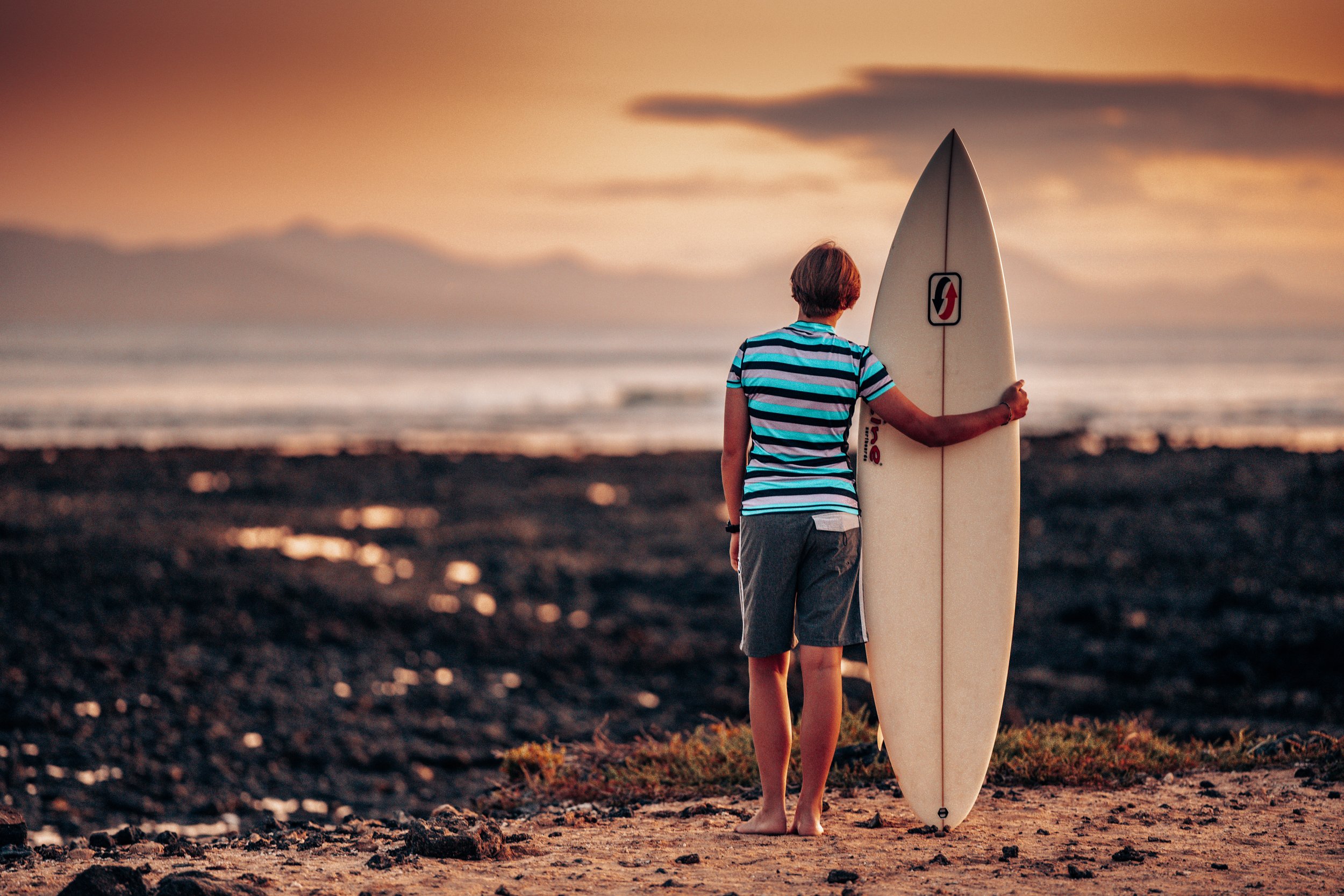 Person standing on a beach at sunset holding a surfboard, facing the ocean.