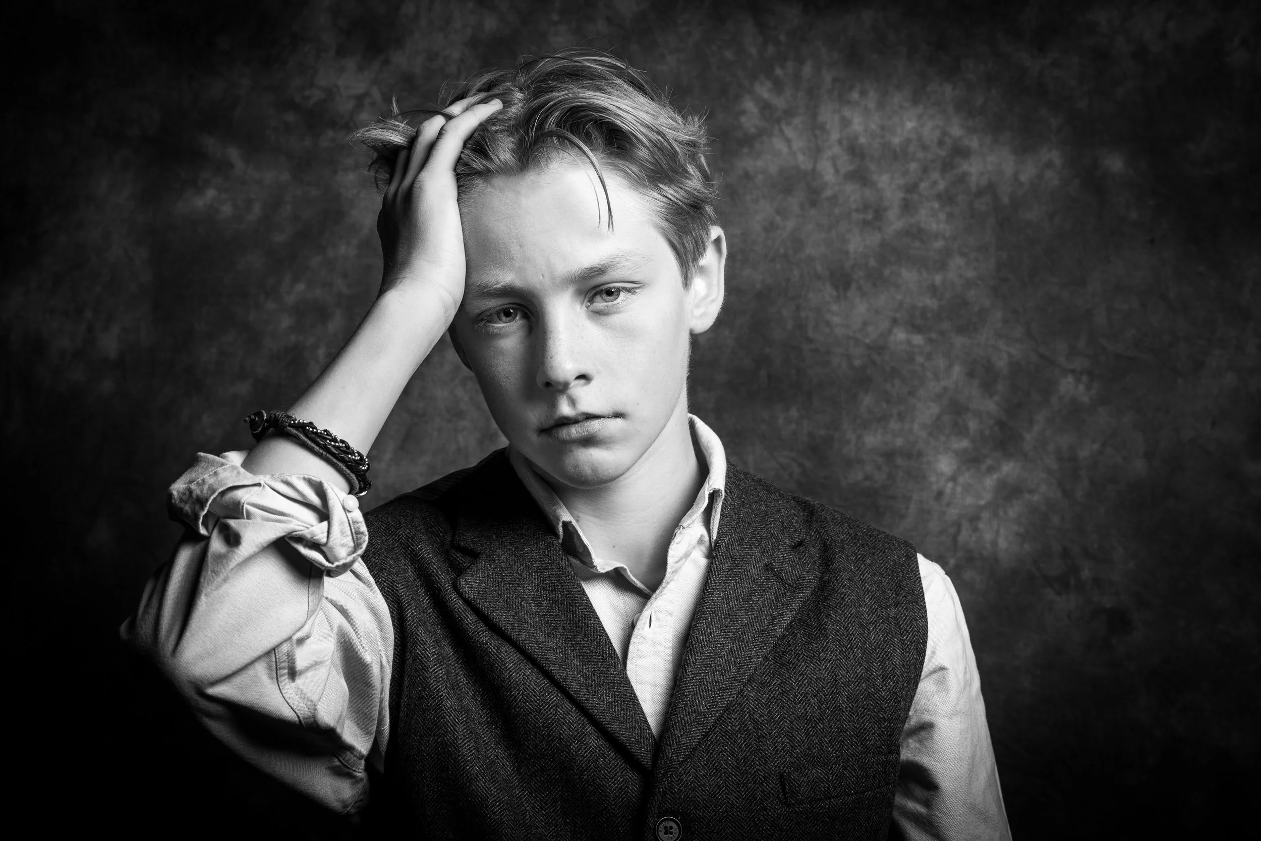 Black and white portrait of a young man with messy hair, wearing a vest over a button-up shirt, with one hand on his forehead, looking serious.