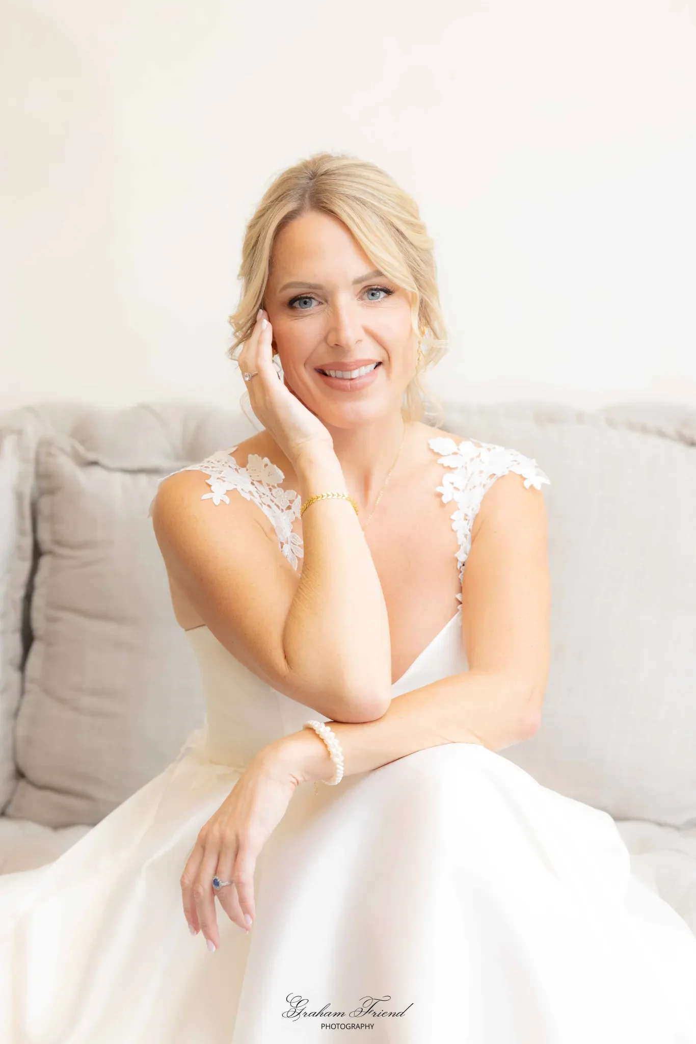 A blonde woman in a white dress with lace shoulder details, smiling, sitting on a beige couch with a neutral background.