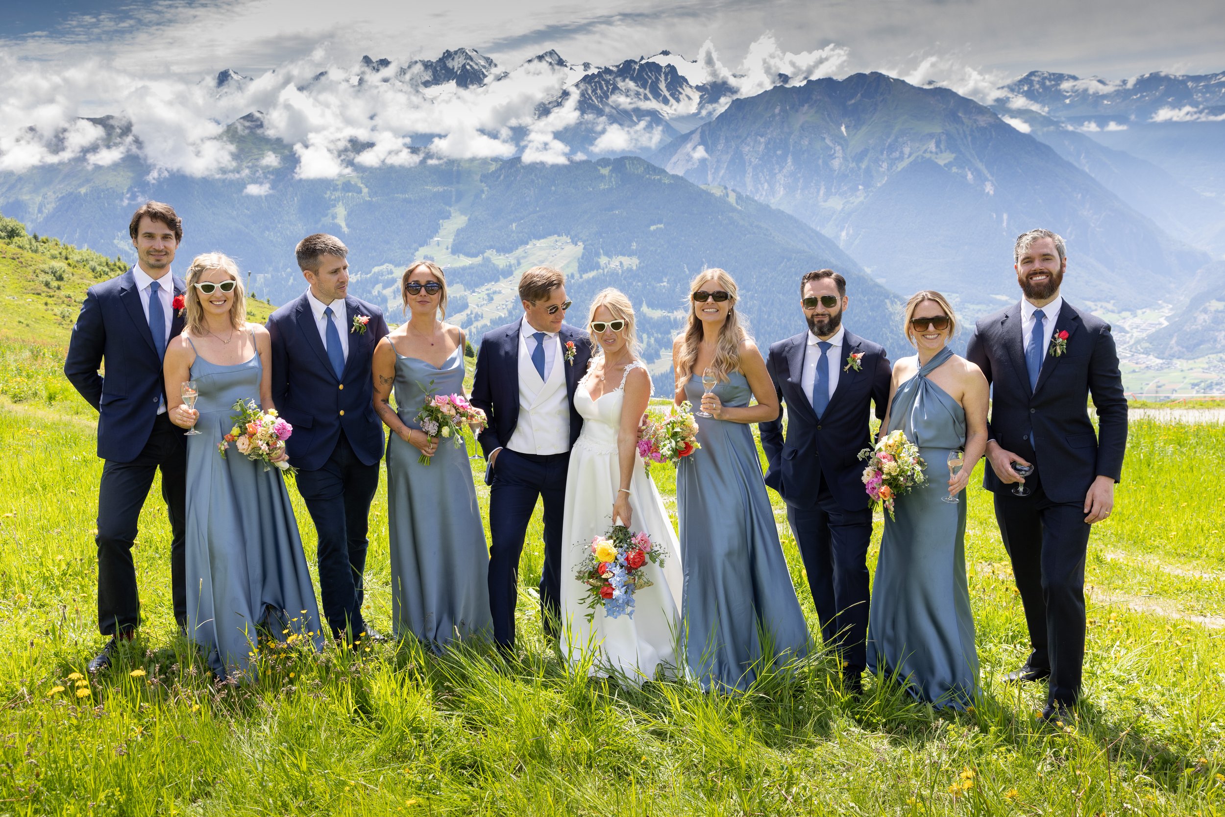 A Verbier Switzerland wedding partyy standing outdoors in a green field with mountains in the background. The group includes bridesmaids in blue dresses, groomsmen in suits, and a bride and groom in the center holding bouquets and champagne glasses.