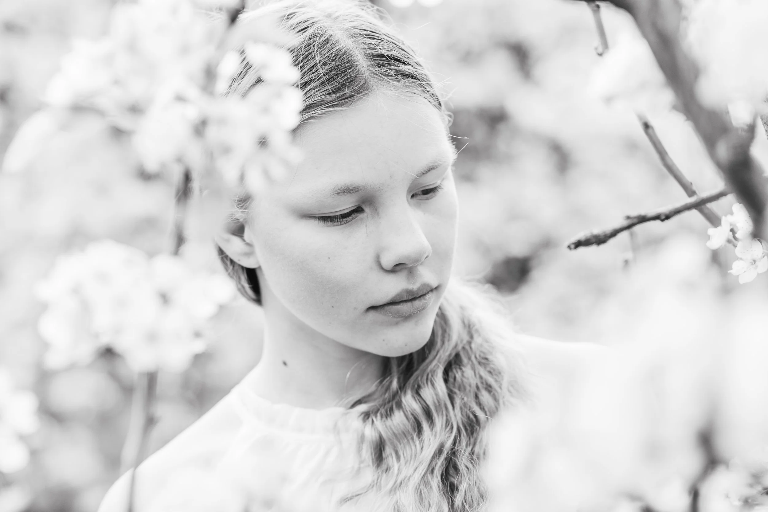 Black and white photo of a young woman with long, wavy hair, looking down amidst flowering branches.