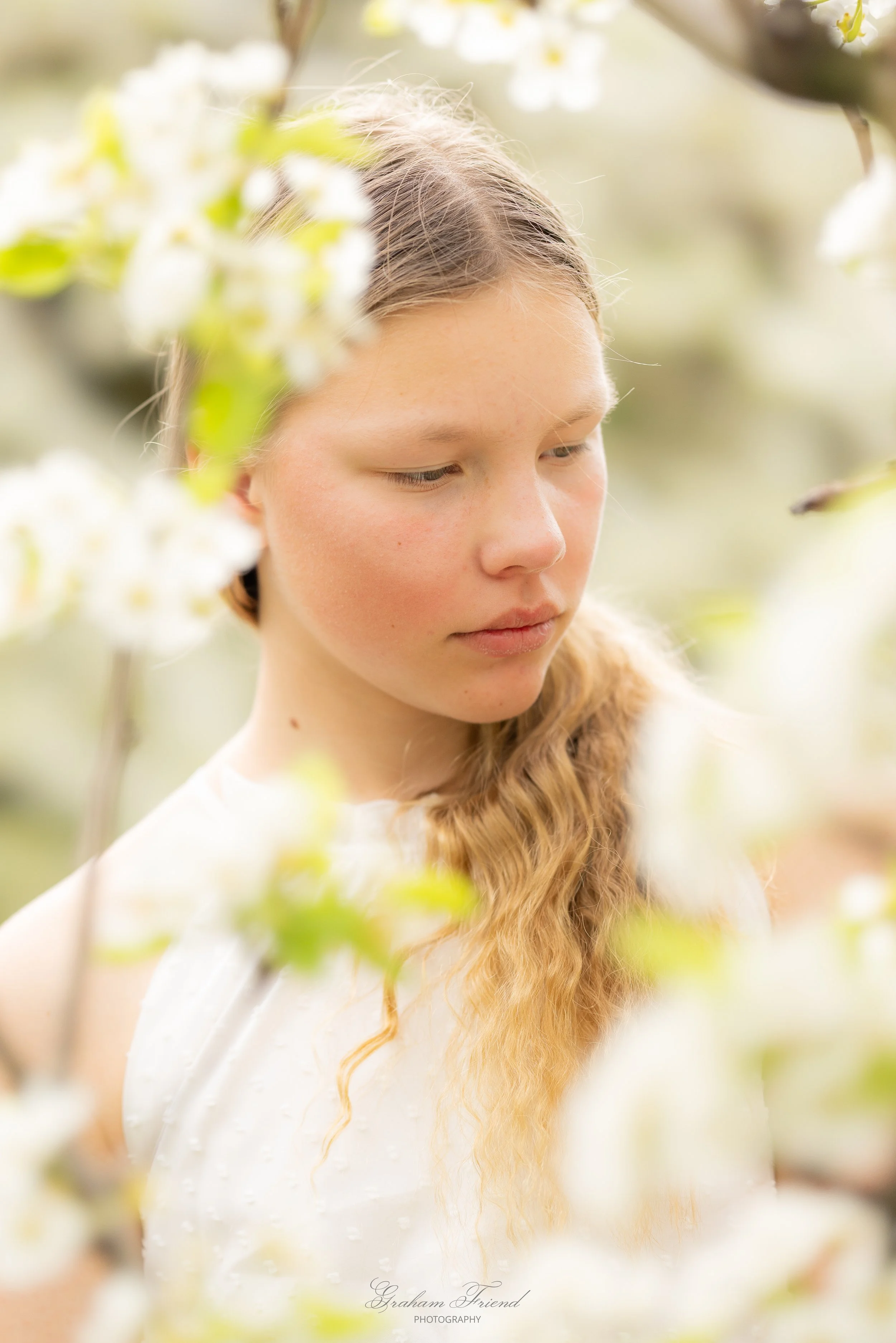 Young woman with long, wavy hair standing among blossoming white flowers, looking down contemplatively in a natural outdoor setting.