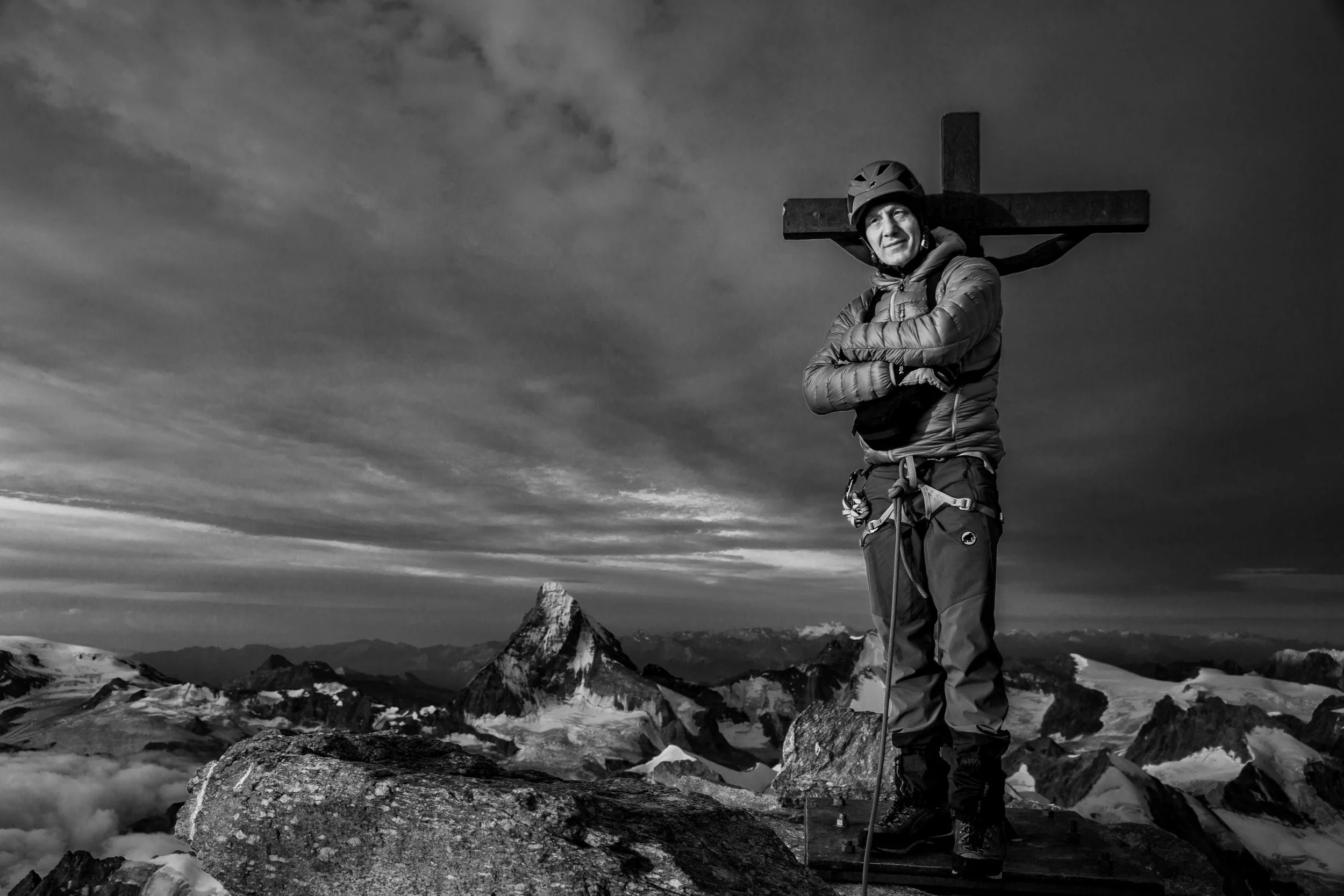 A person standing on a mountain peak at sunset with a cross on their back, in a black and white photograph.