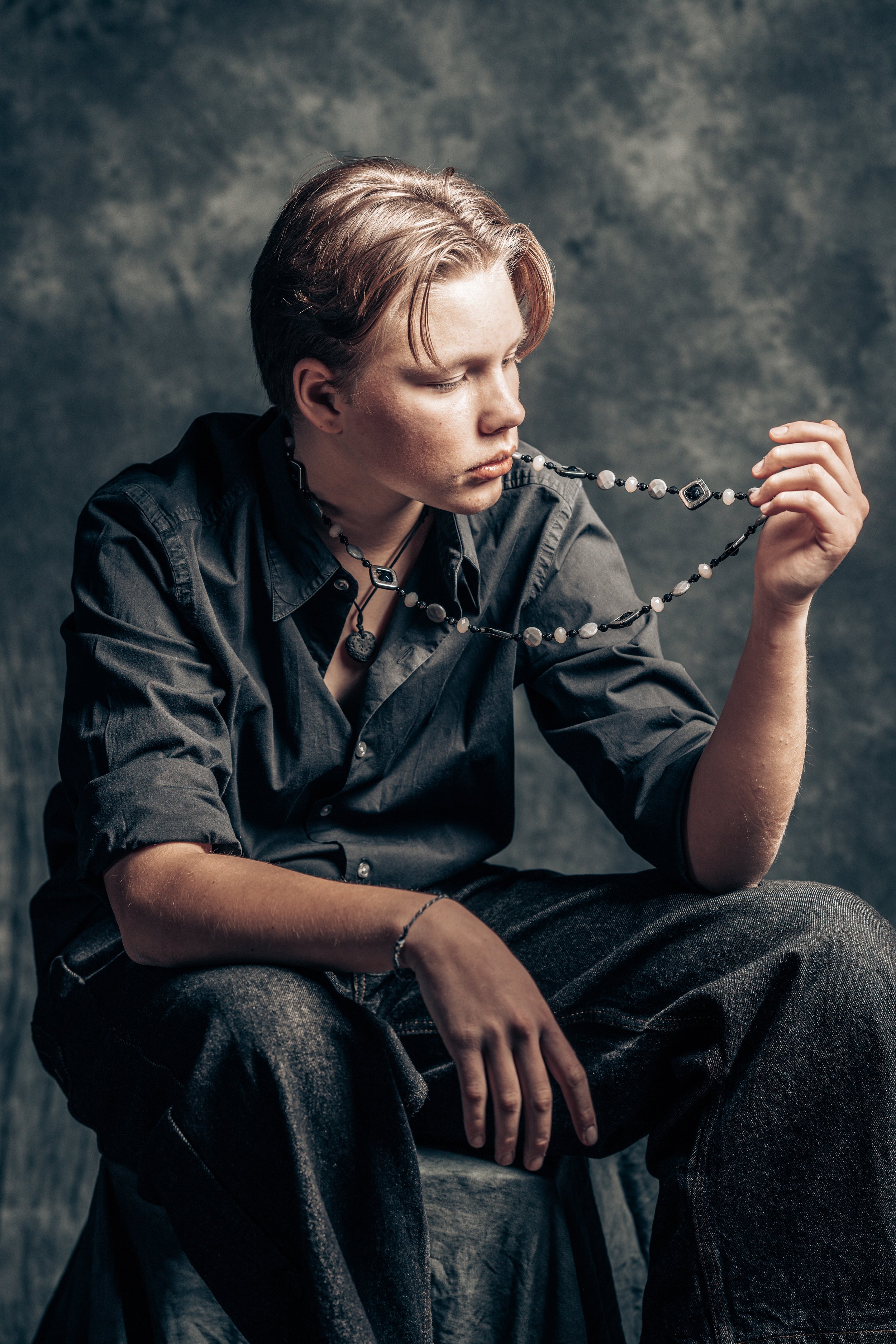 A young person with short, light brown hair, wearing a black button-up shirt, sitting with one leg crossed over the other, looking down and holding a beaded necklace close to their mouth against a dark, textured background.