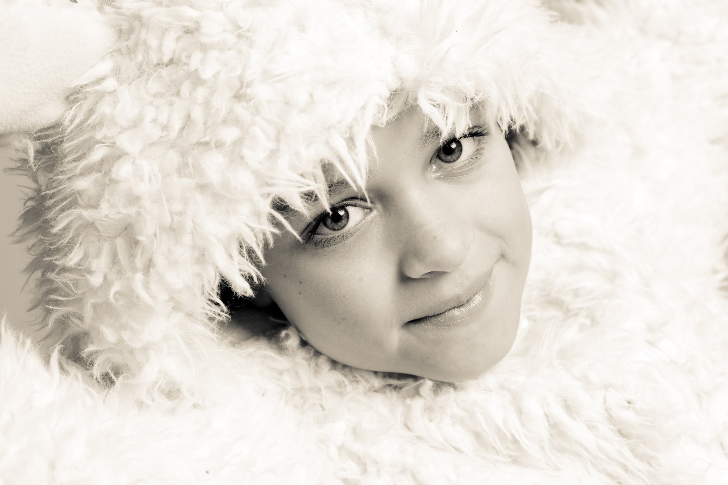 Close-up of a smiling child's face, wrapped in a fluffy, fuzzy white blanket or garment, with bright eyes and soft facial features.