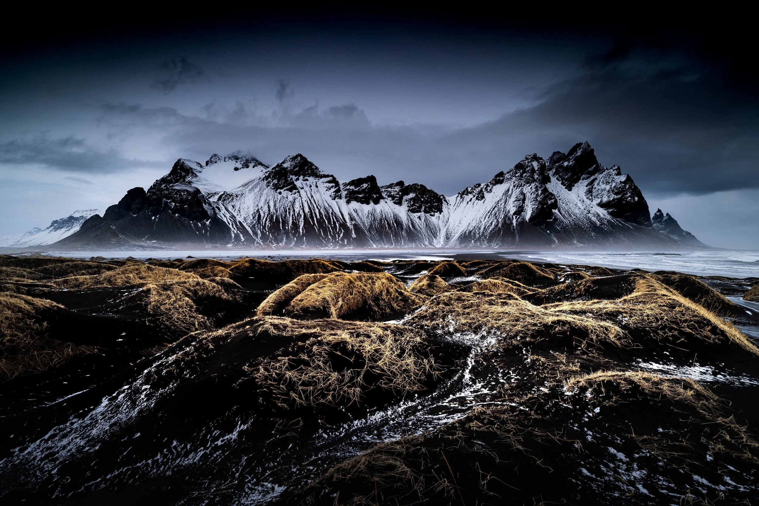 Snow-capped mountains under cloudy sky with patches of dry grass on dark volcanic soil in the foreground.