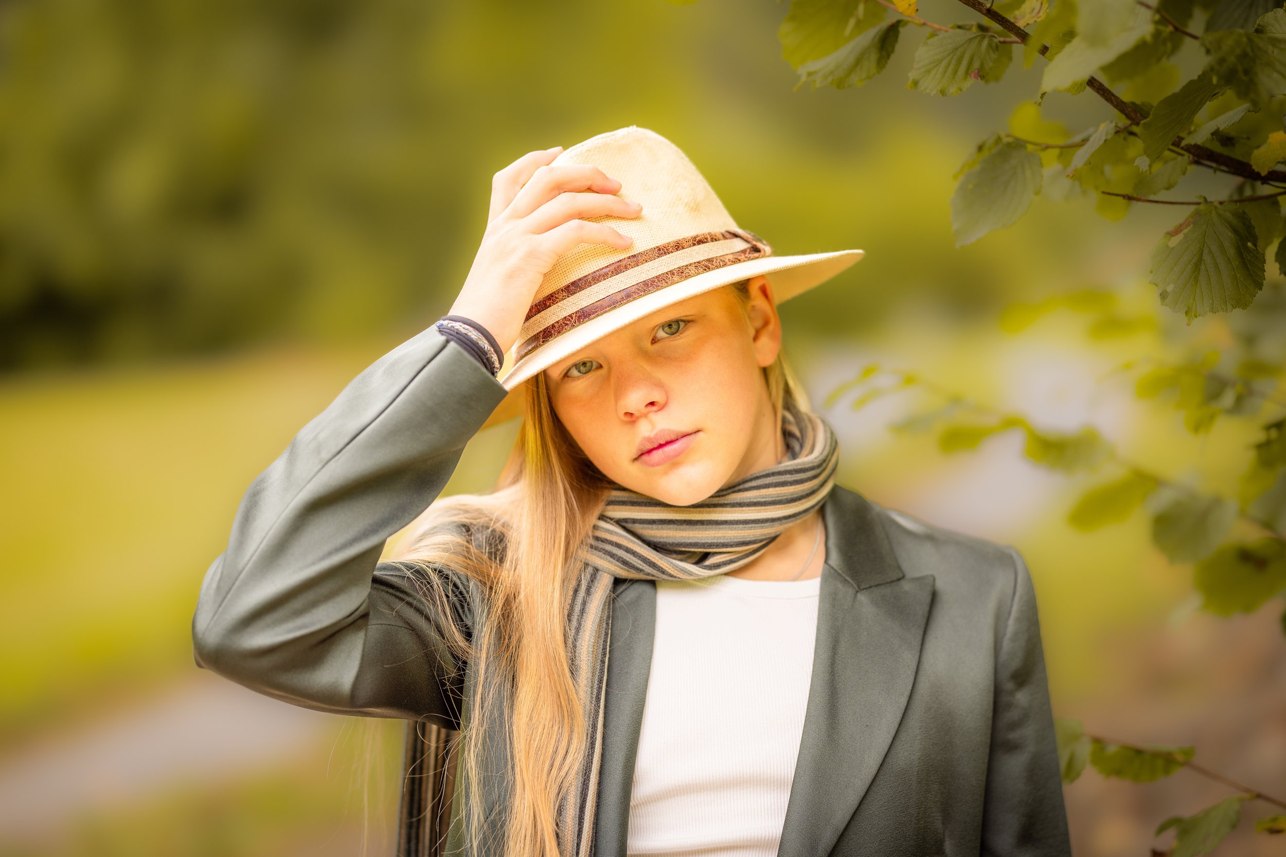 A young woman with long blonde hair wearing a beige fedora hat, gray blazer, striped scarf, and white shirt, standing outdoors with green foliage background, tilting her head and touching her hat.