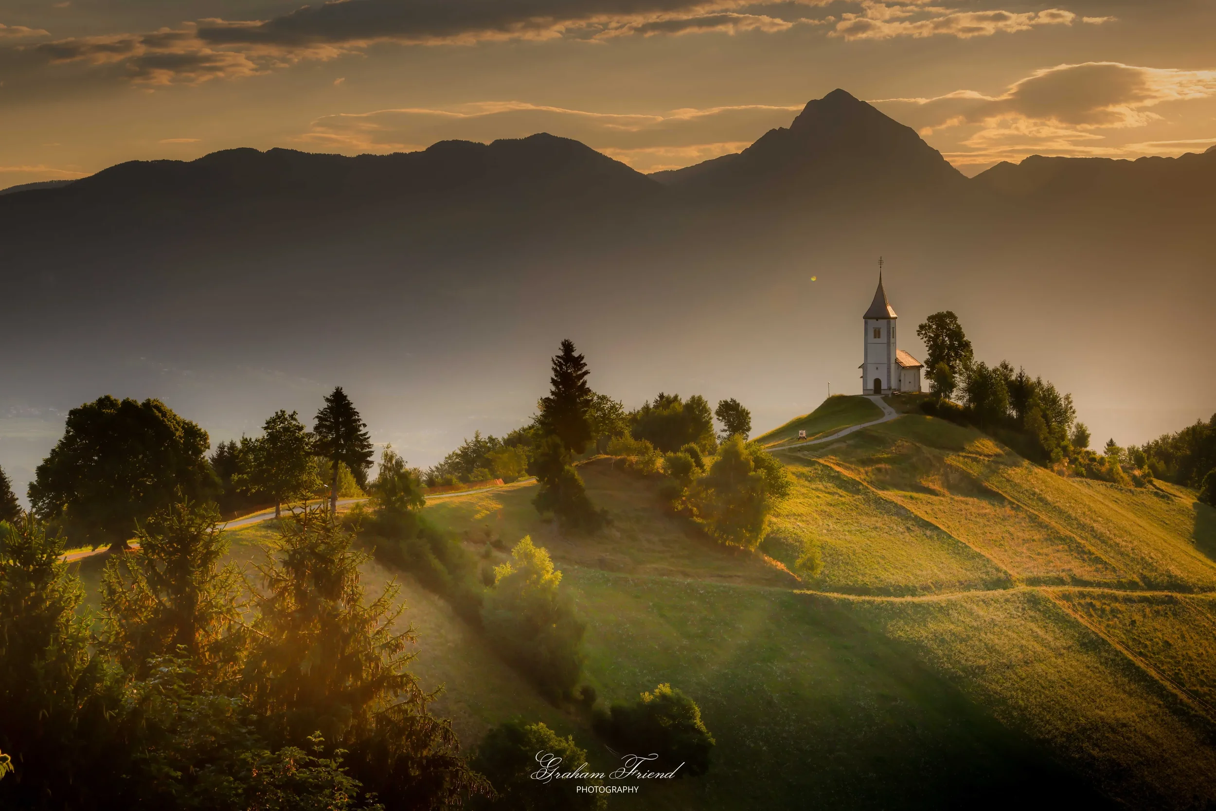 Sunset over a small hill with a church and trees, mountains in the background, and a misty sky.