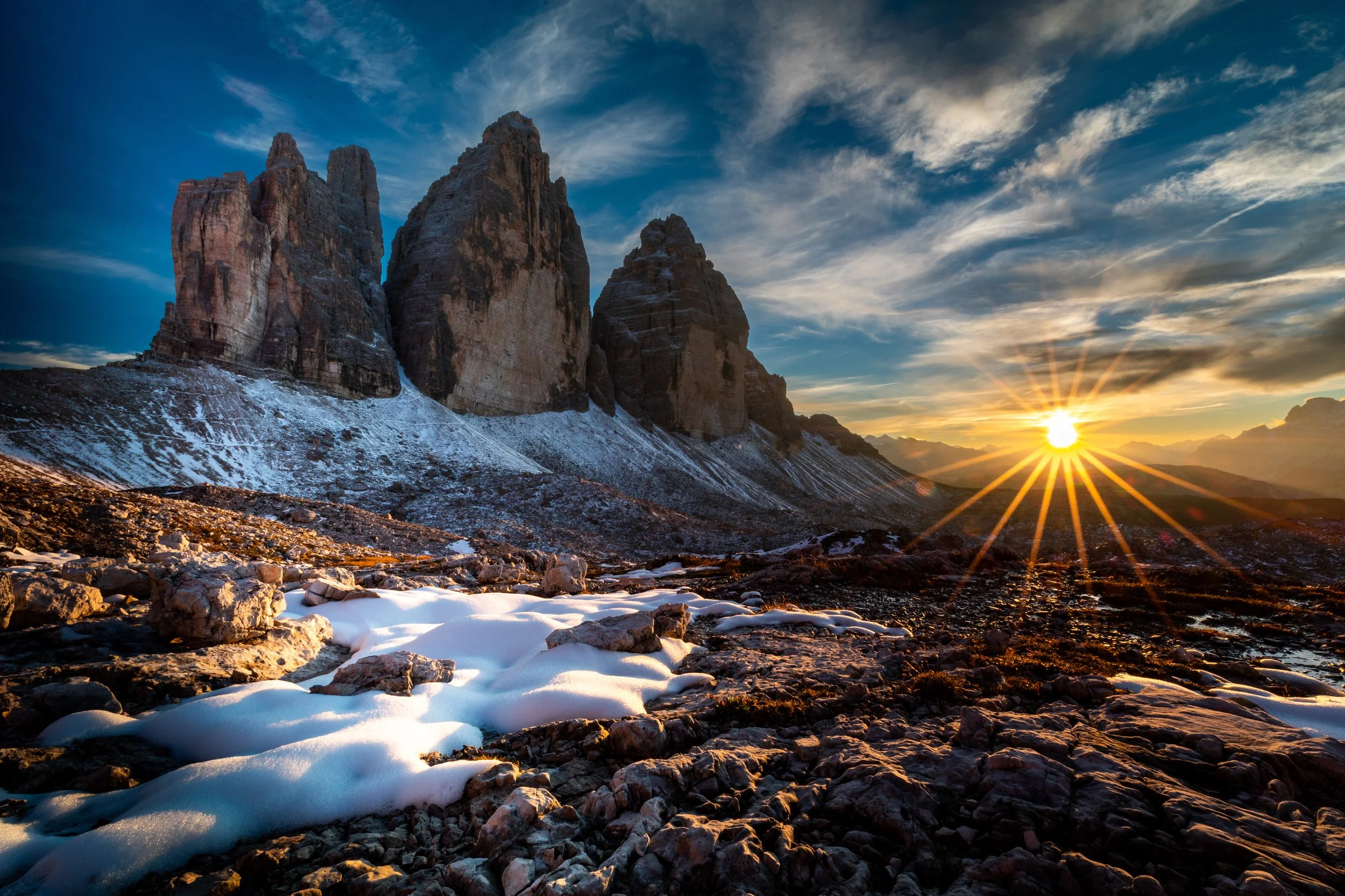 Sunset over a rocky, snow-covered mountain landscape with three large mountain peaks and a partly cloudy sky.