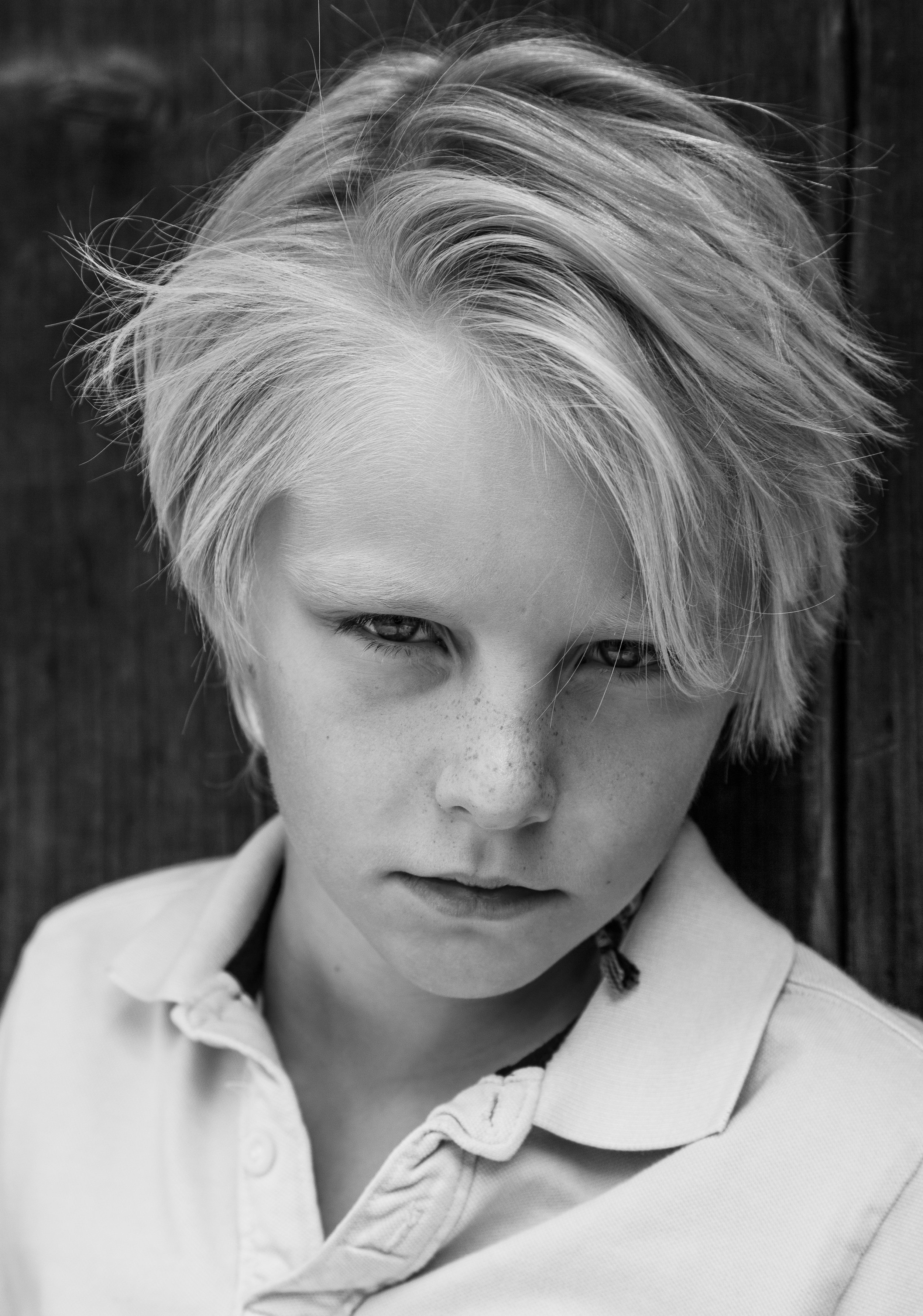 Close-up black and white portrait of a young boy with tousled blond hair and light skin, wearing a collared shirt, against a wooden background.