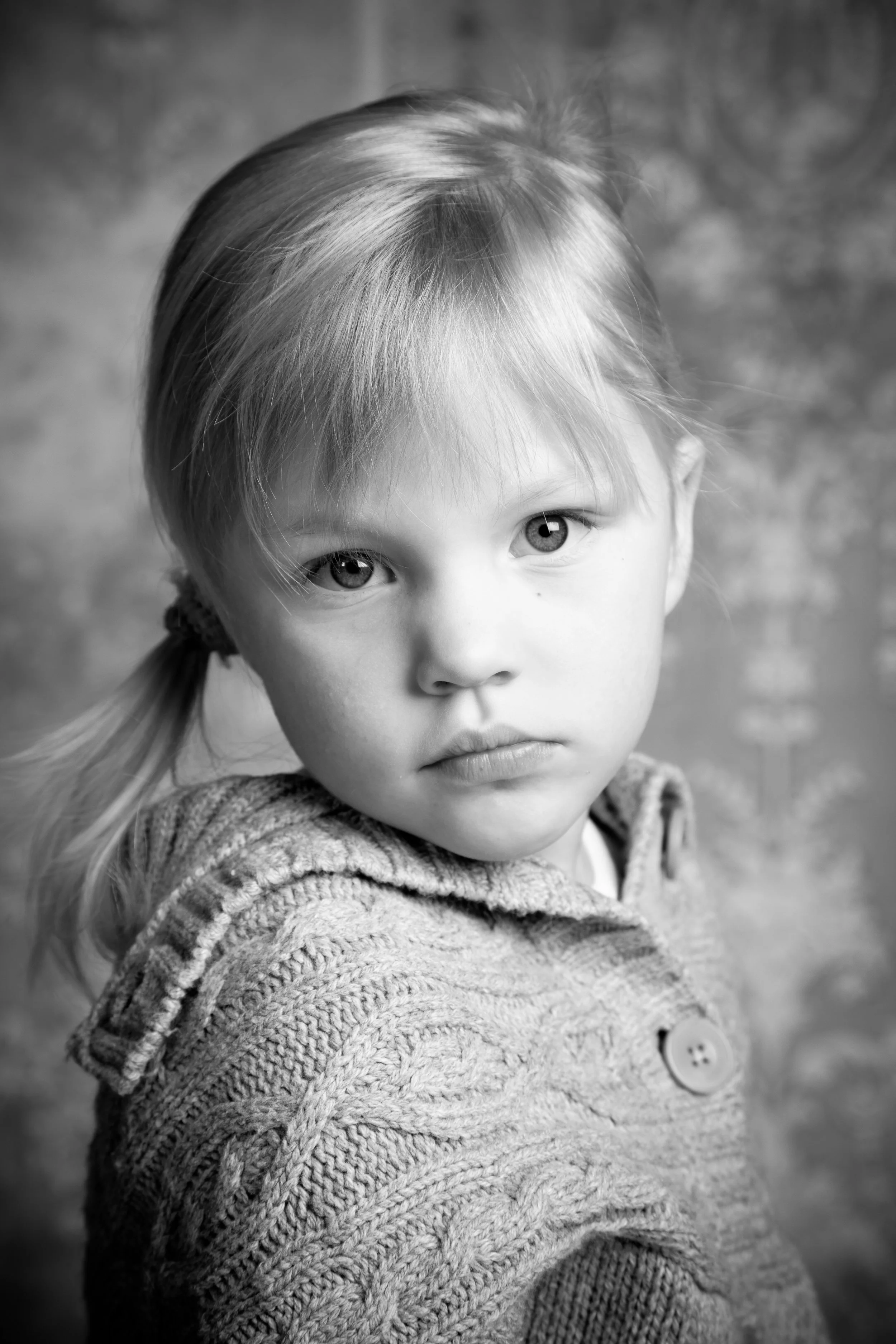 A black-and-white portrait of a young girl with light-colored hair, styled in pigtails, wearing a textured knitted sweater with buttons, looking directly at the camera with a serious expression.
