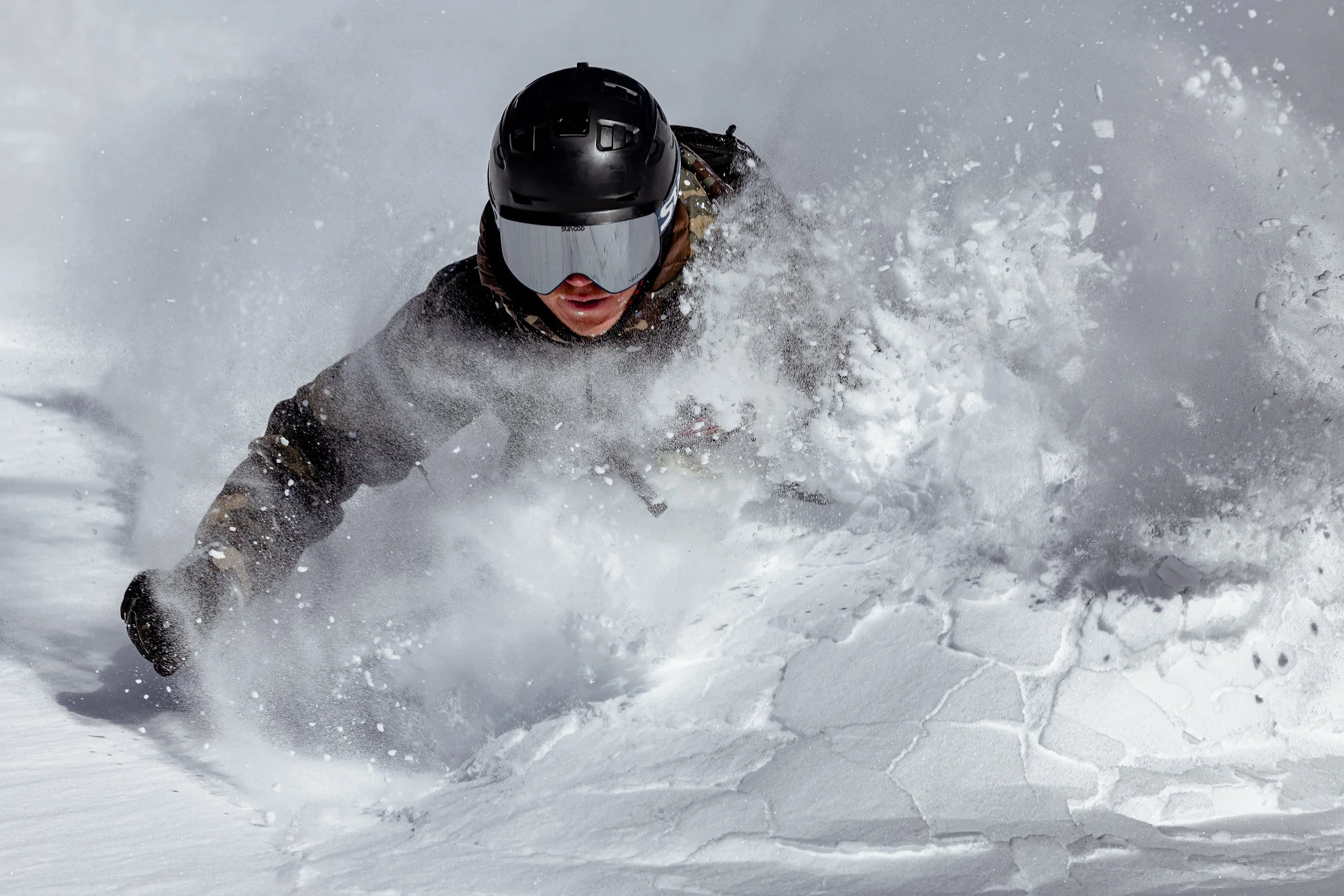 Person wearing a black helmet and ski goggles, skiing down a snowy slope, kicking up snow.