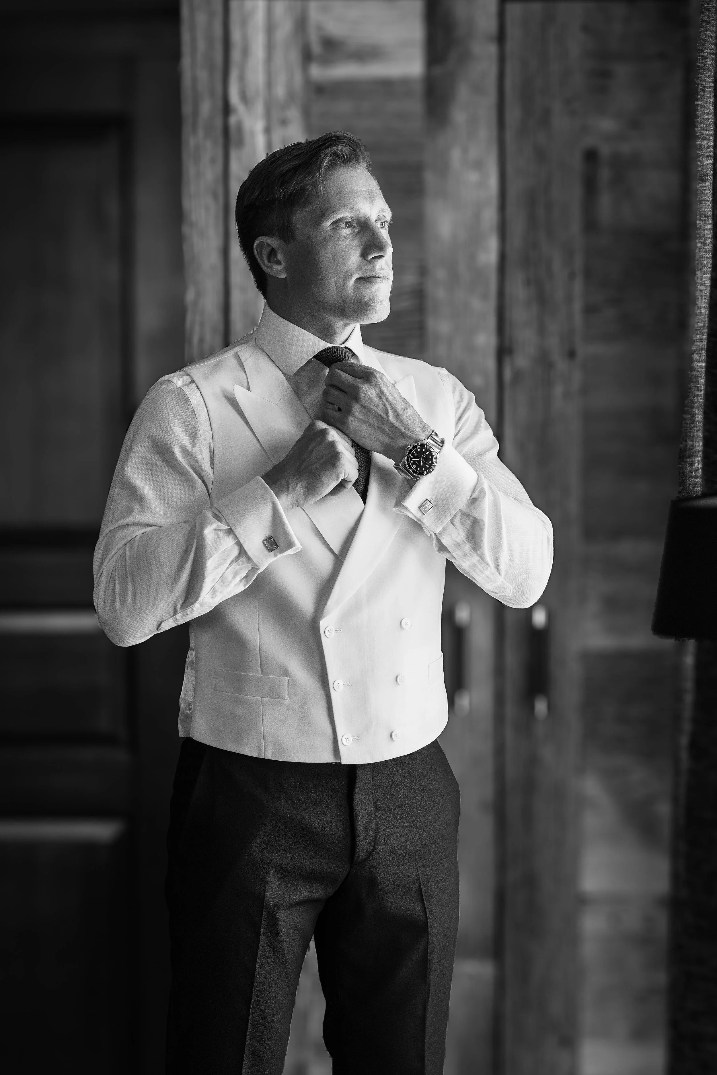 A man in a tuxedo adjusting his tie in a wooden interior setting, black and white photo taken in The Farinet Hotel, Verbier, Switzerland.